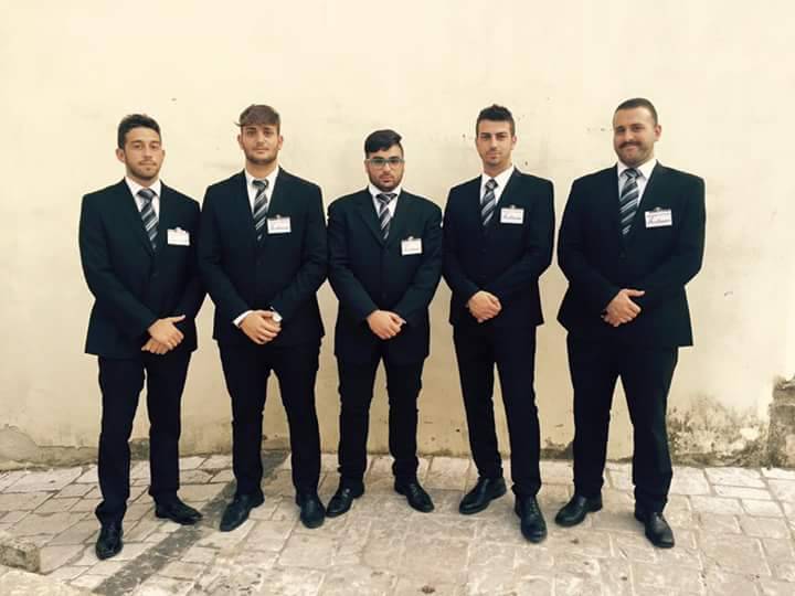 Cinque uomini in giacca e cravatta nera sono in piedi davanti a un muro chiaro, ognuno con un cartellino con il suo nome.