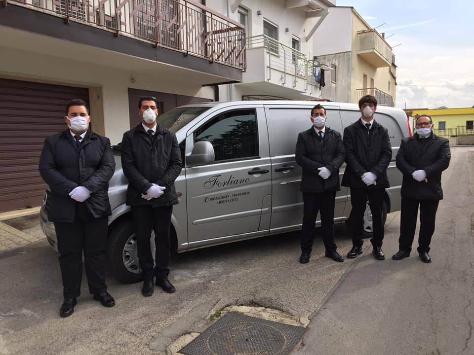 Cinque persone in abiti neri, maschere e guanti sono in piedi vicino a un carro funebre argentato in strada.