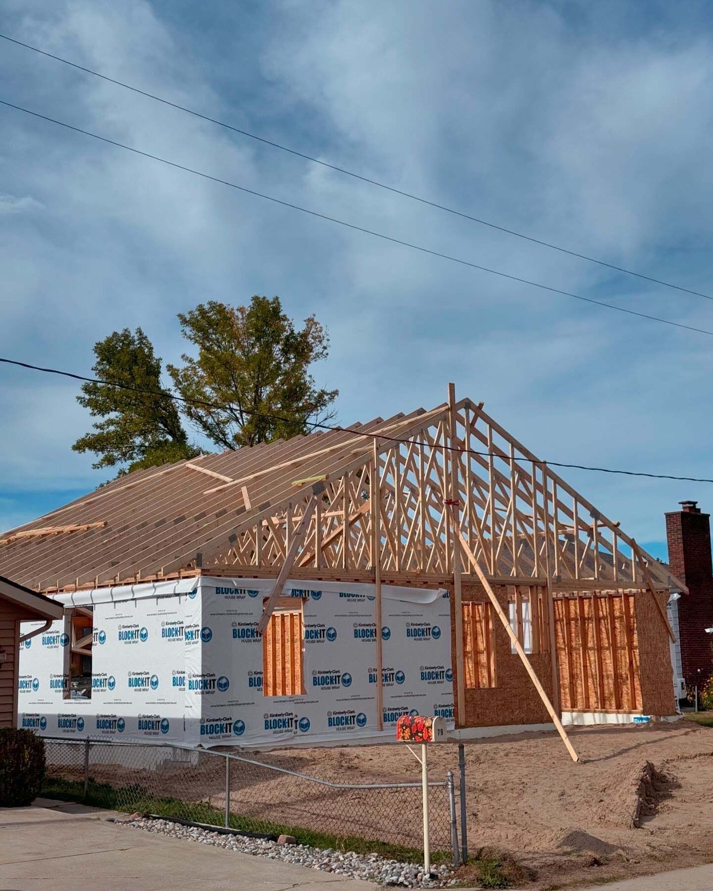House under construction; wooden frame, sheathing, and blue and white wrap visible against a cloudy sky.