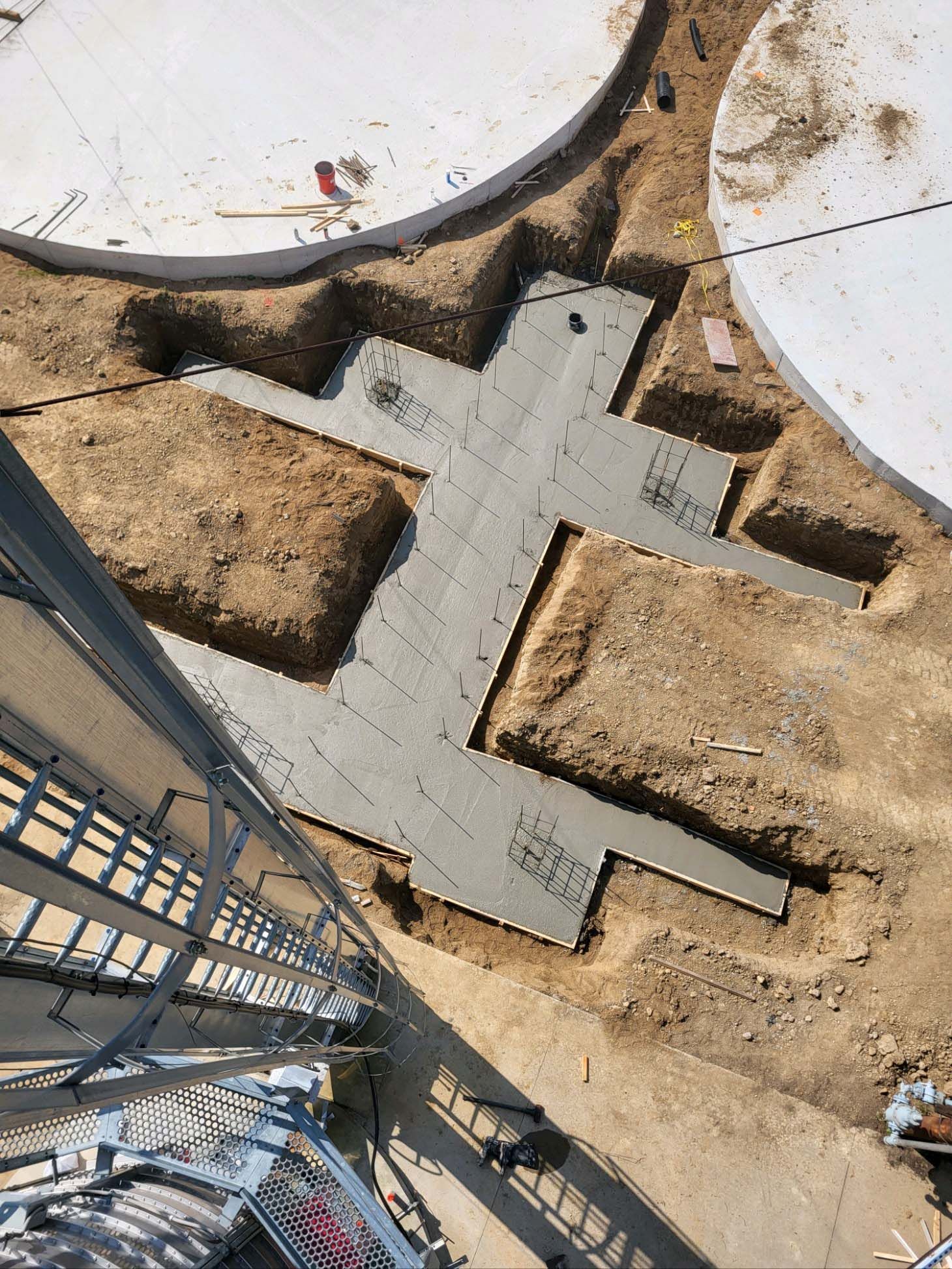 Construction site: recently poured gray concrete foundation between circular structures, surrounded by dirt.