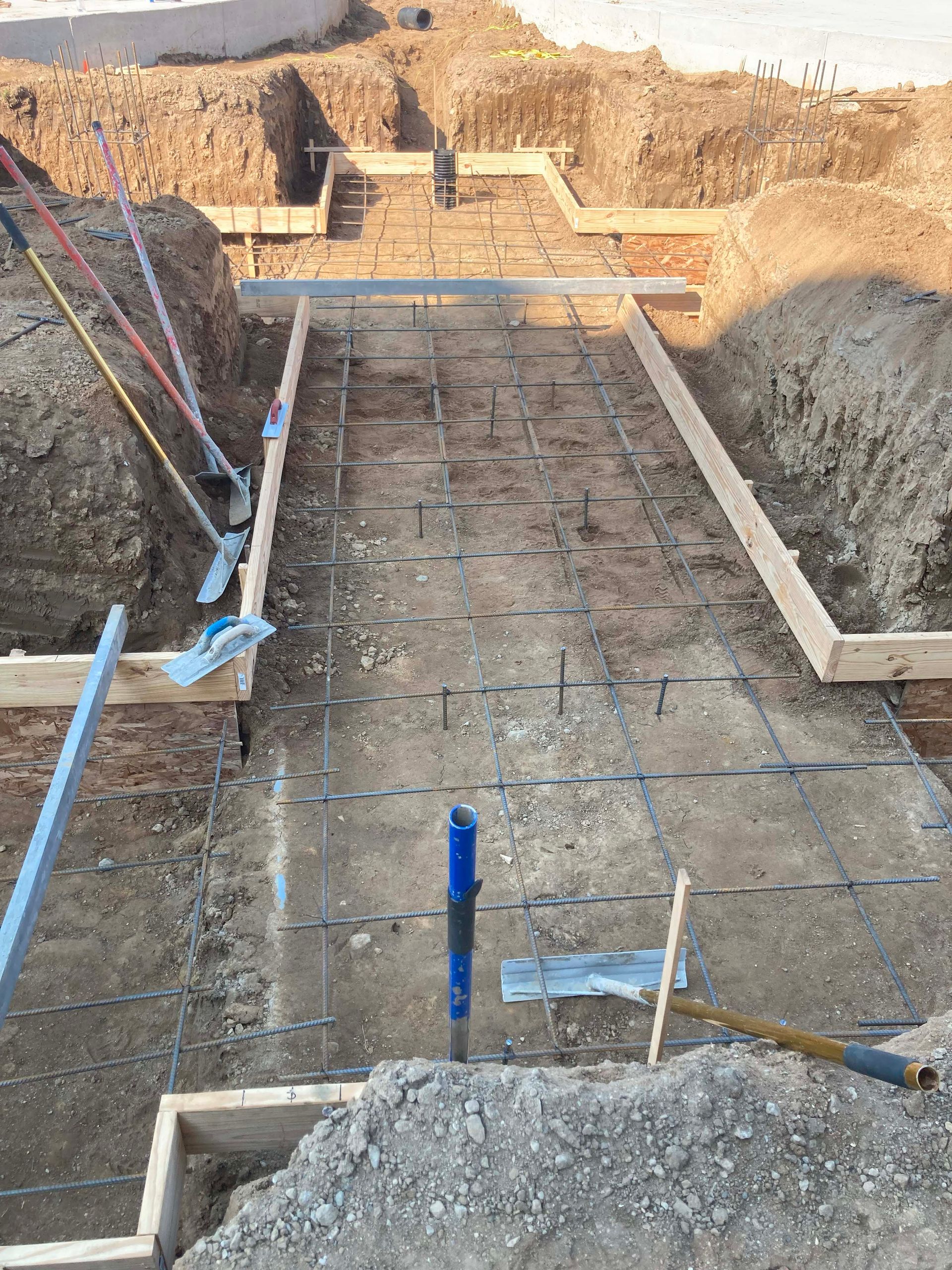 Construction site: excavation with rebar grid in trench, wooden forms, tools laid out.