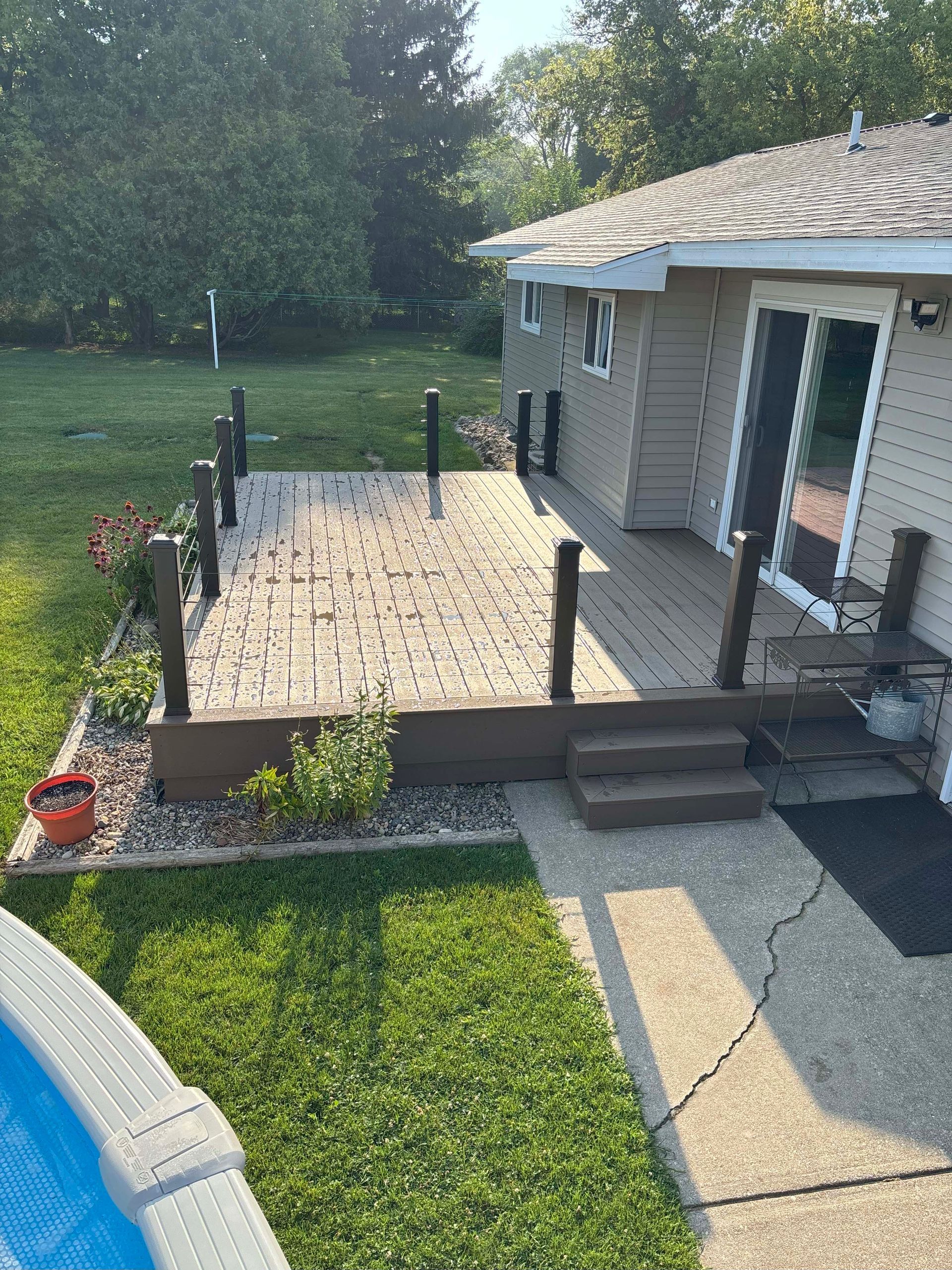 A backyard deck with brown railings, steps, and a stone-like surface next to a house and pool.