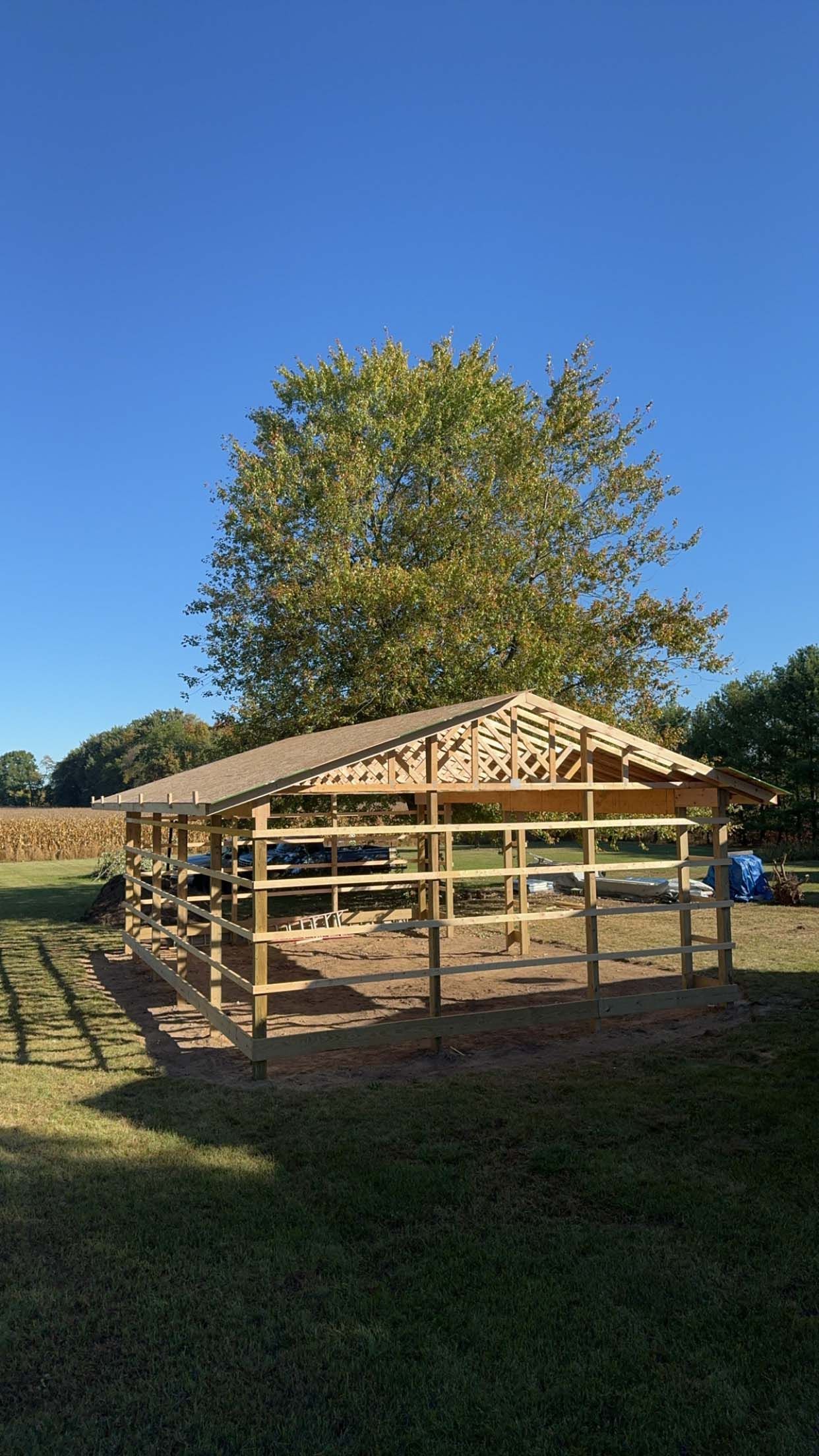Wooden horse shelter with surrounding fence under a blue sky, tree in background.