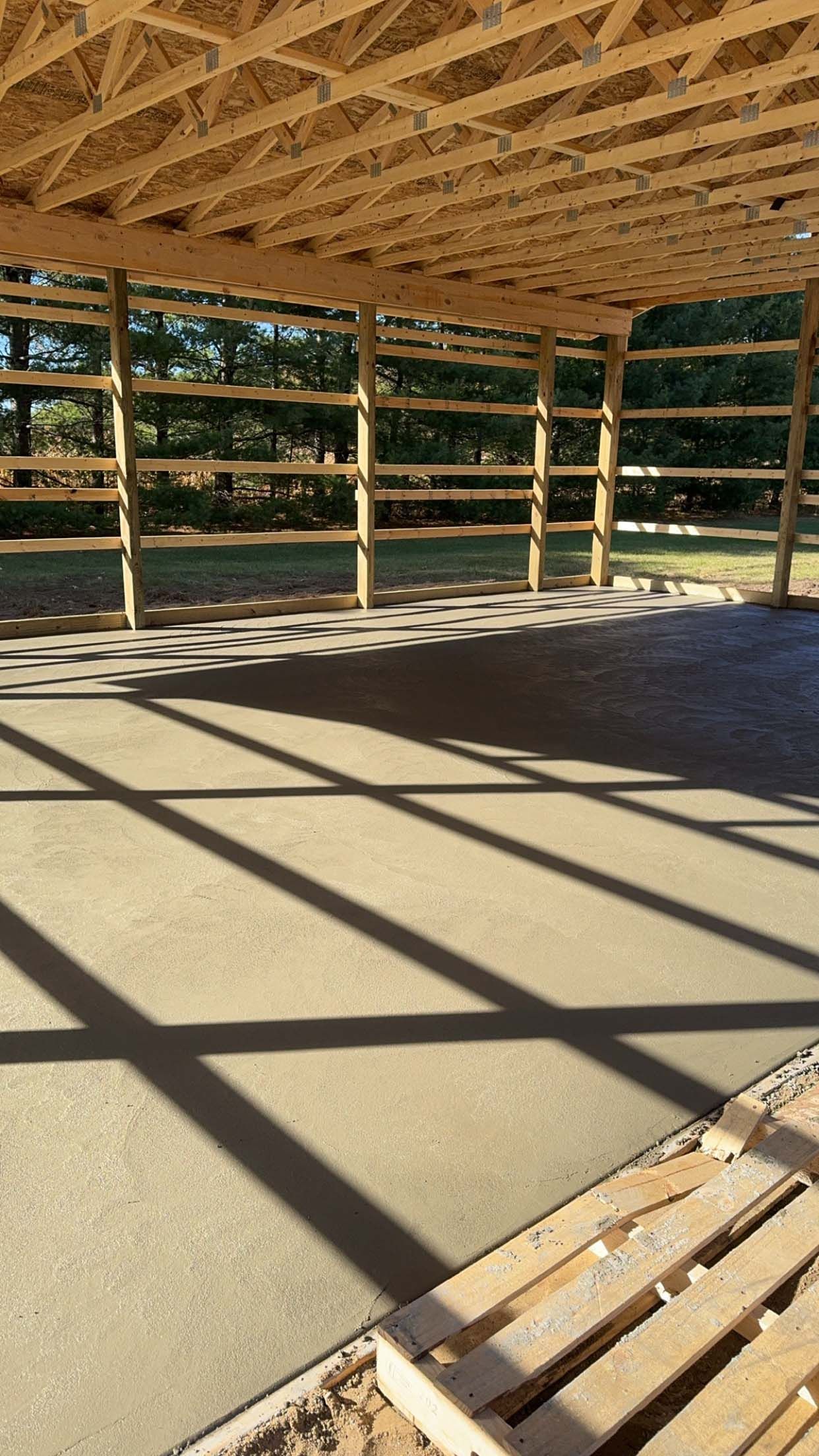 A partially built wooden structure with a concrete floor, casting shadows from the beams above.