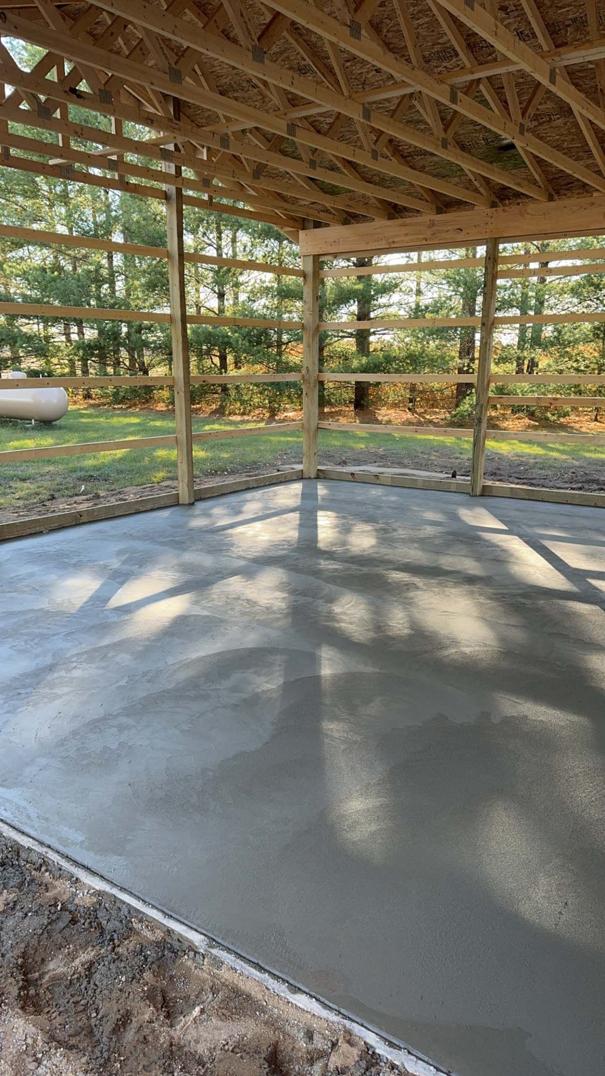 Newly poured concrete floor under a wooden roof structure, set in a grassy outdoor area.