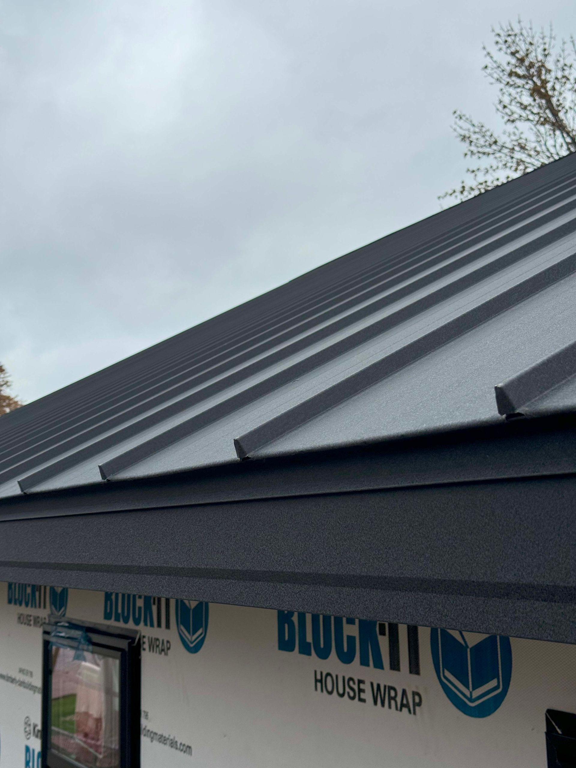Black metal roof on a building with blue house wrap, under a cloudy sky.
