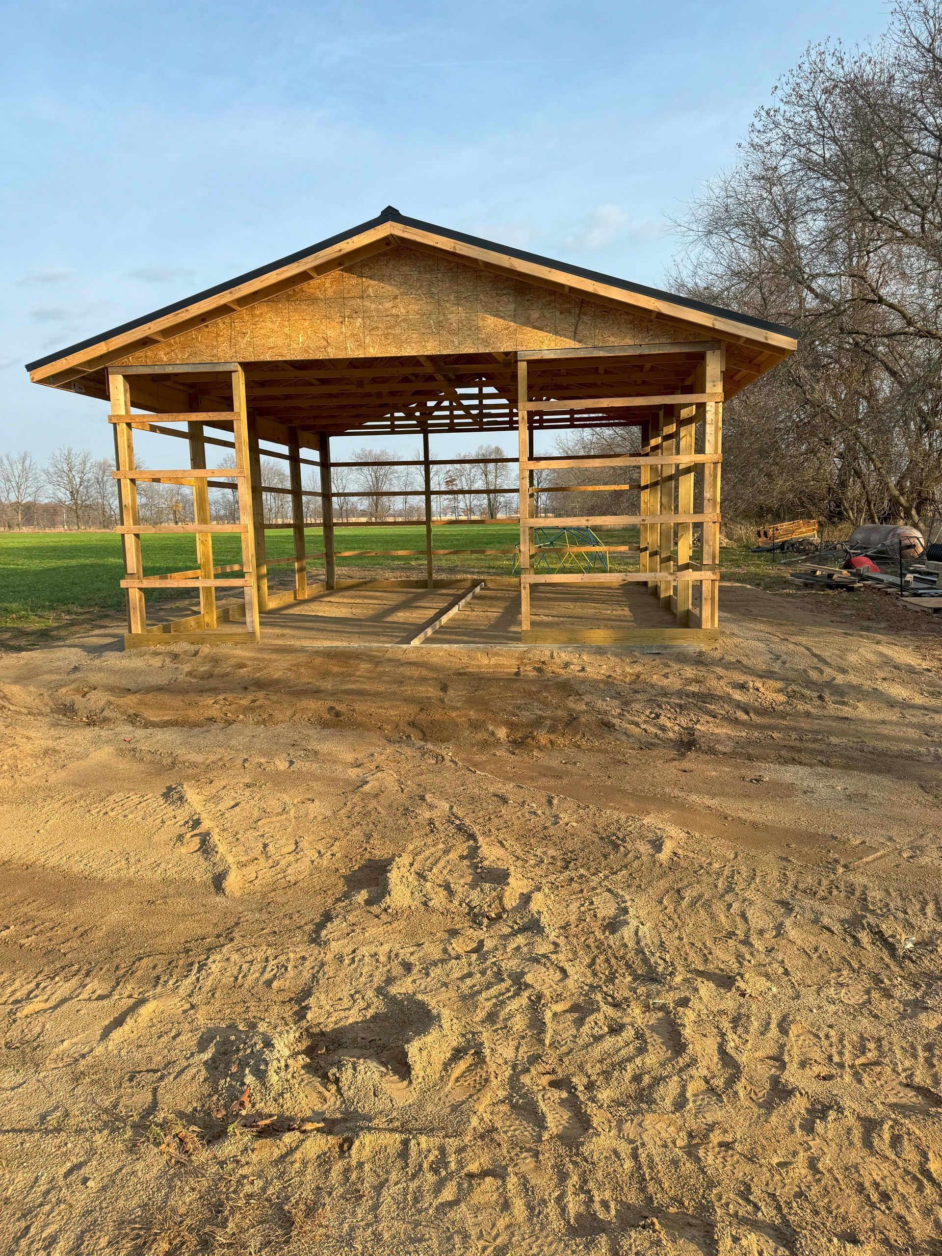 A partially built wooden shed in a field. Brown lumber with a black roof, set against a blue sky.