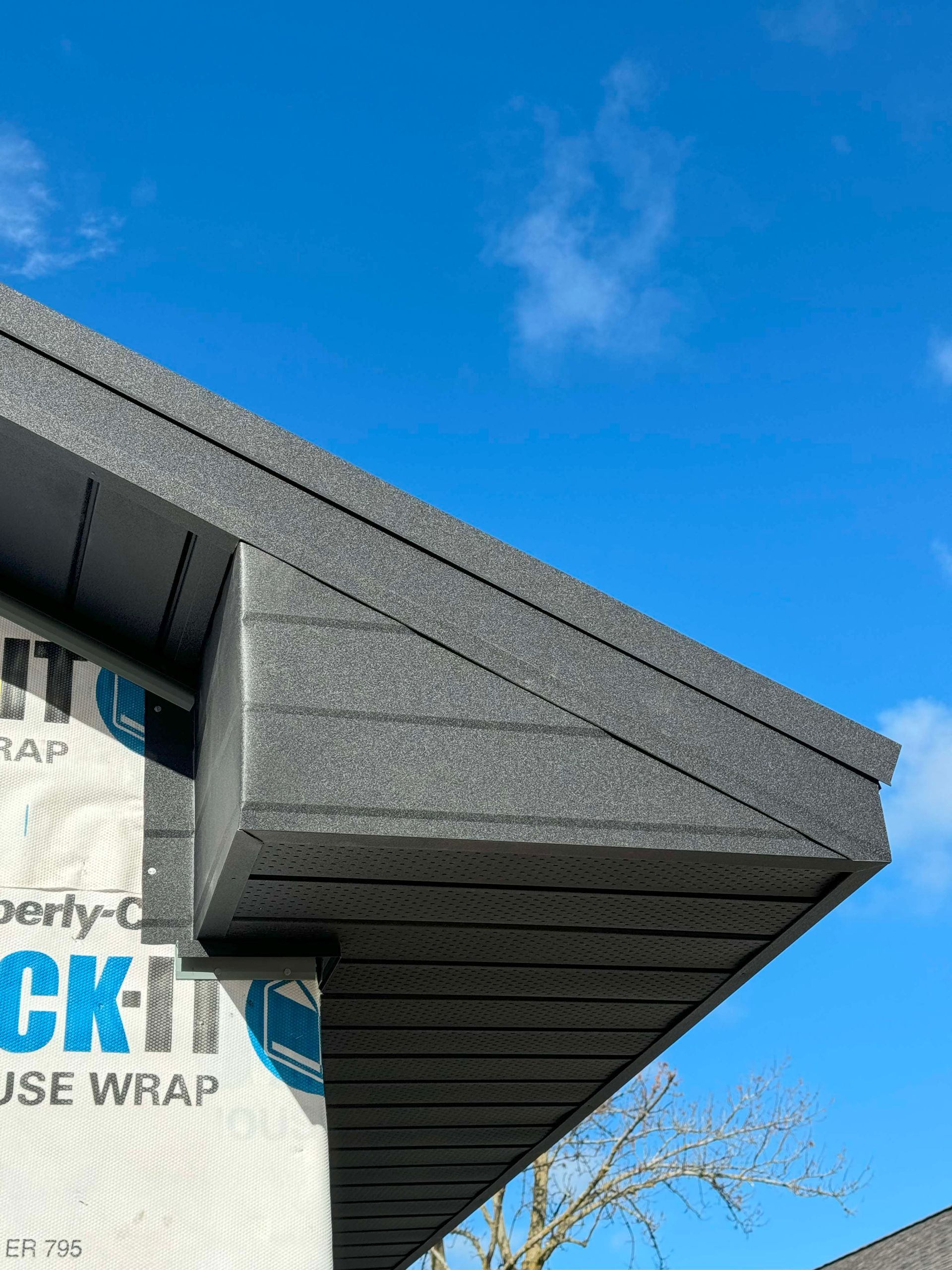 Close-up of a house roof's edge with dark siding, white house wrap, and a blue sky.