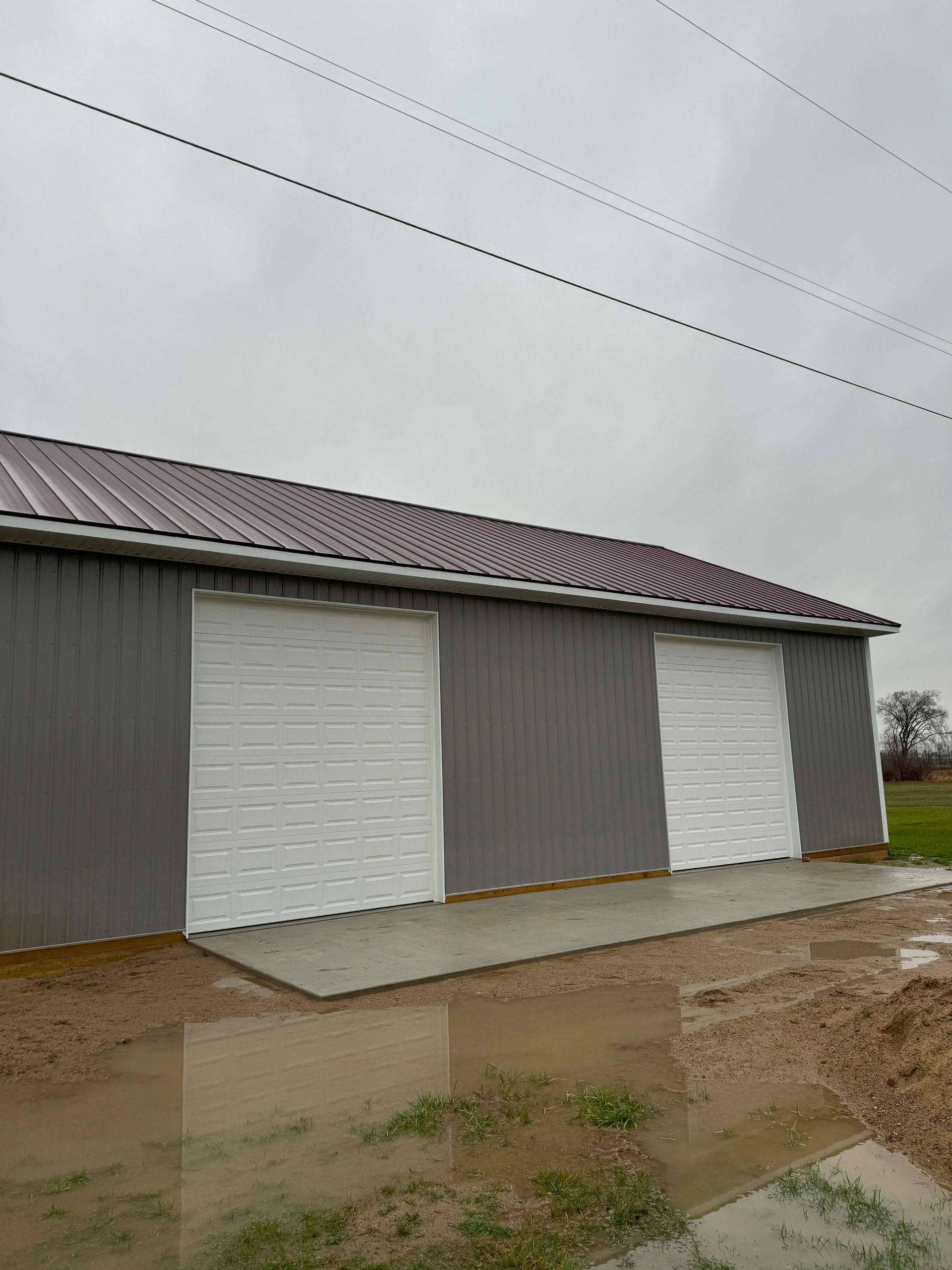 Gray metal building with three white garage doors and a burgundy roof. Overcast day with standing water.