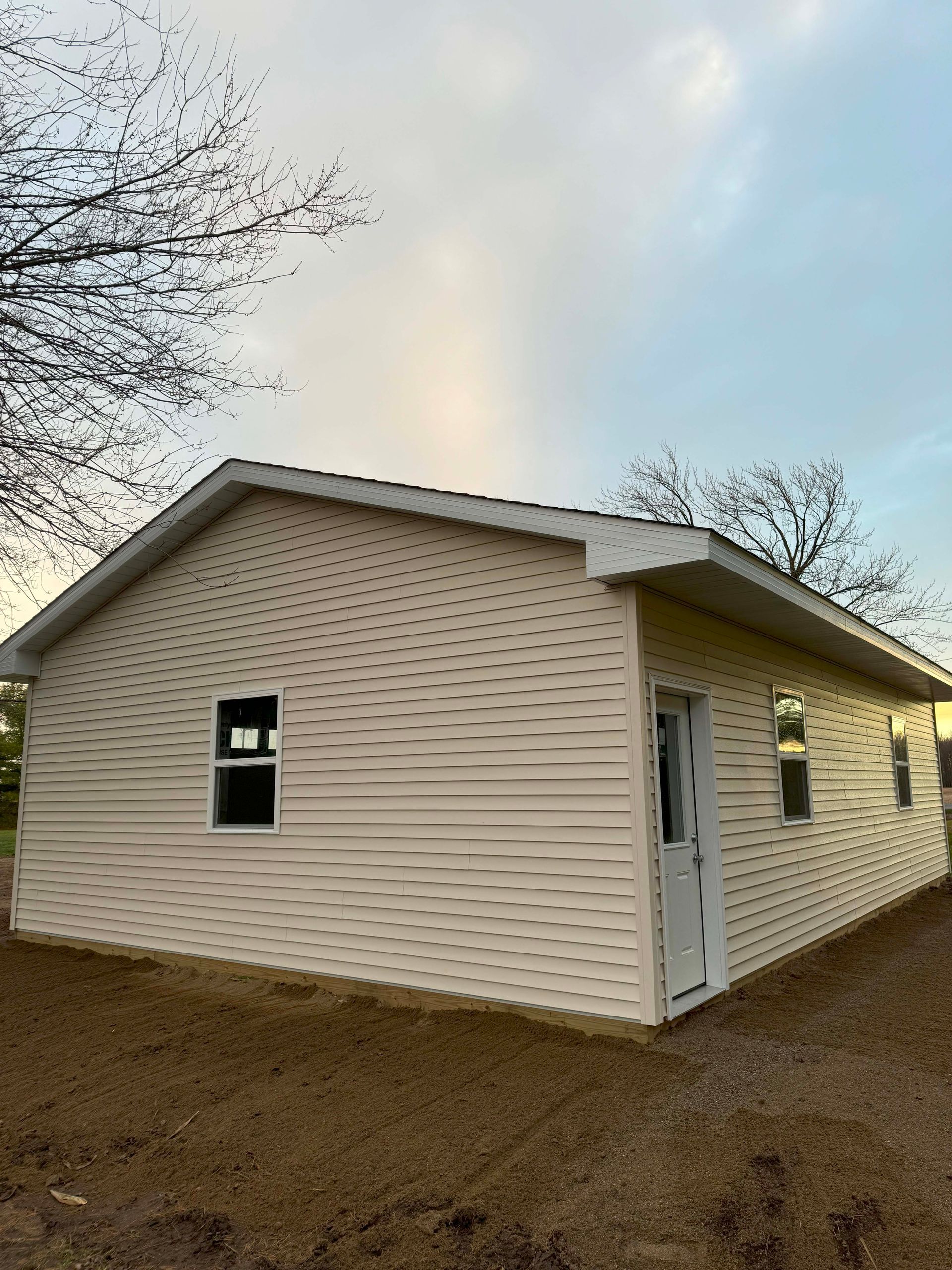 Beige-sided rectangular building with white trim, windows, and door. Set outdoors, under a cloudy sky.