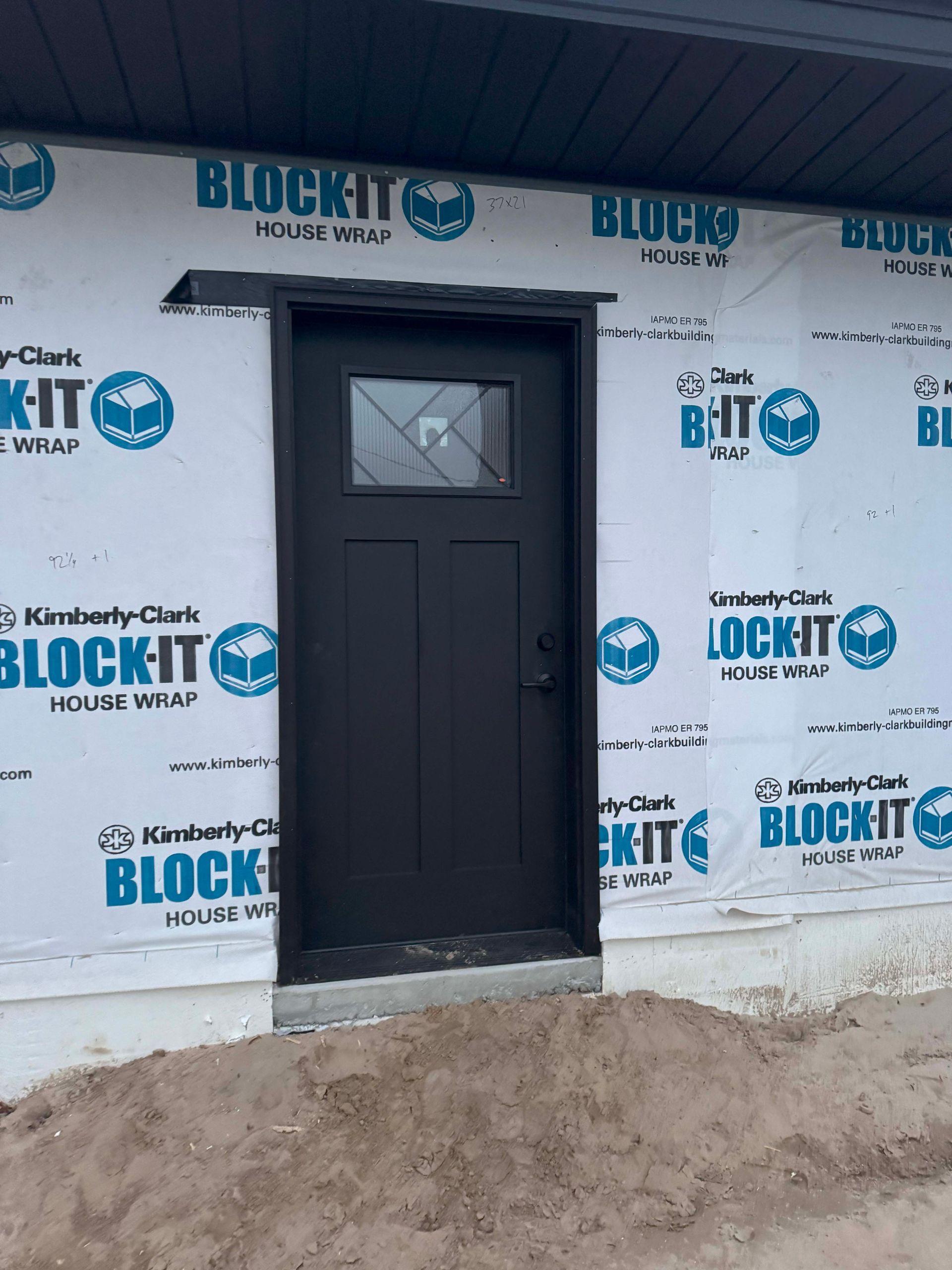 Black front door with a window under a small overhang; surrounded by blue house wrap.