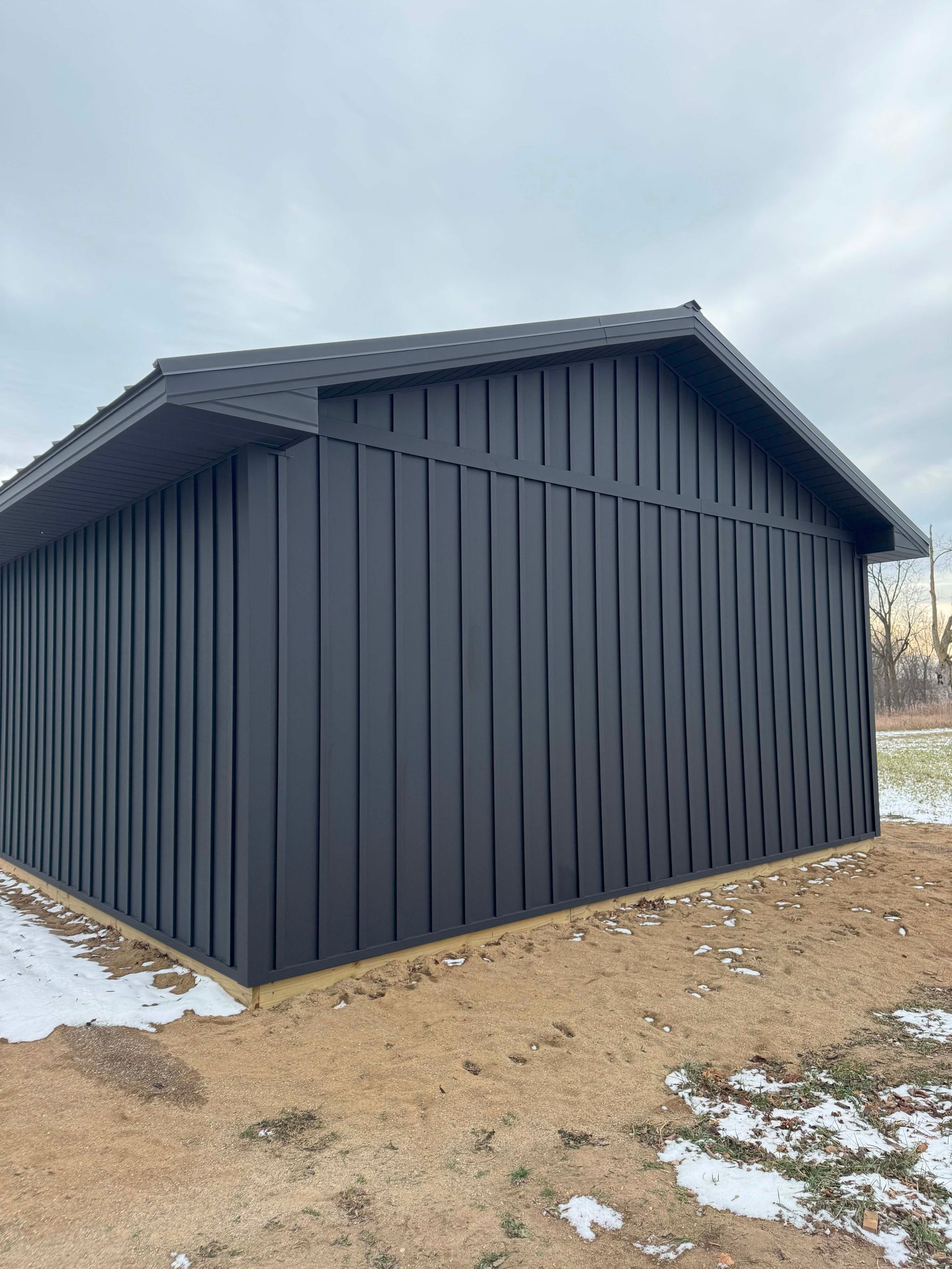 Black vertical-paneled building with dark shingle roof, on a gravel and snow-covered ground.