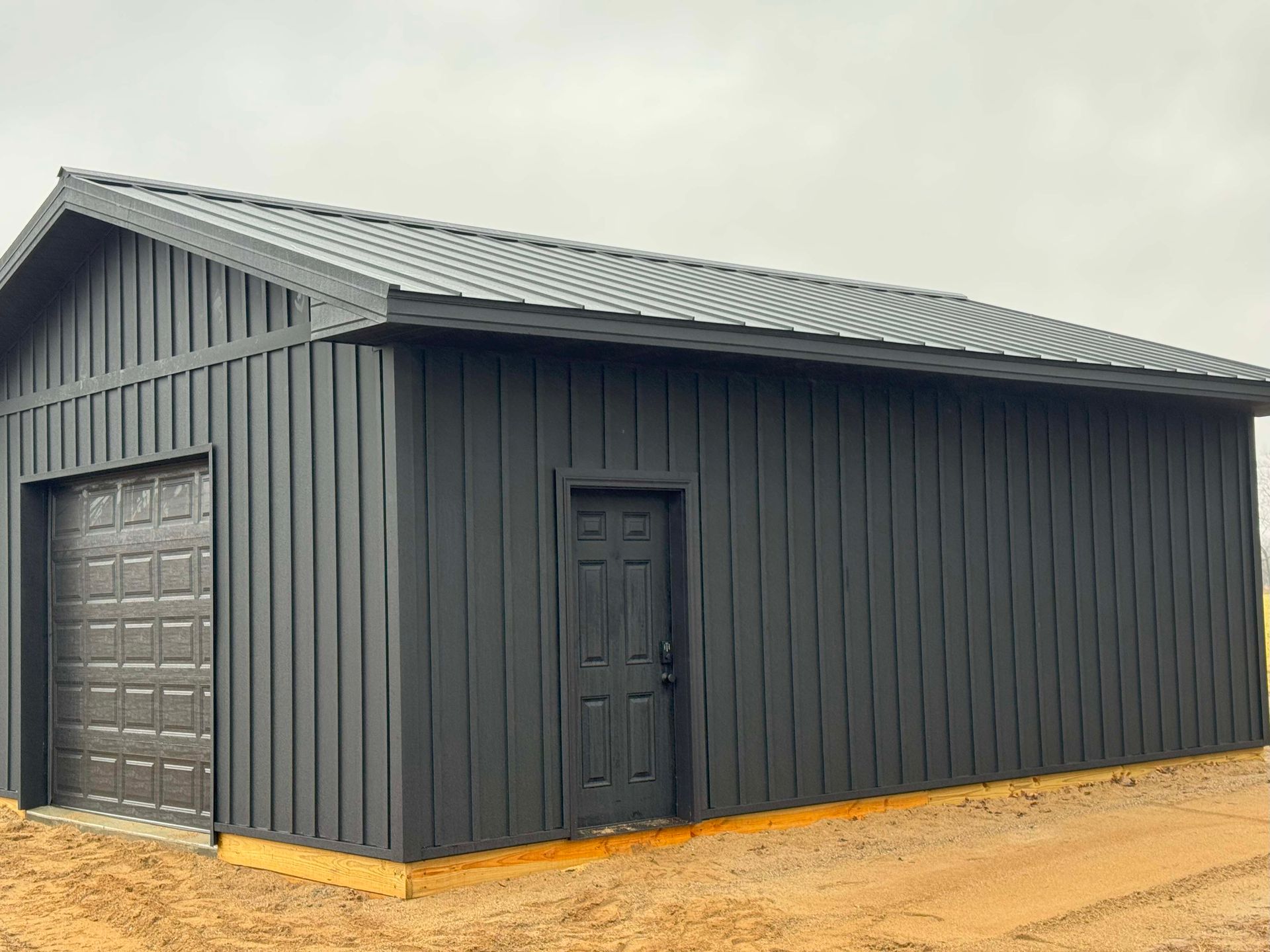 Black metal-roofed garage with a black door and siding; set on dirt.