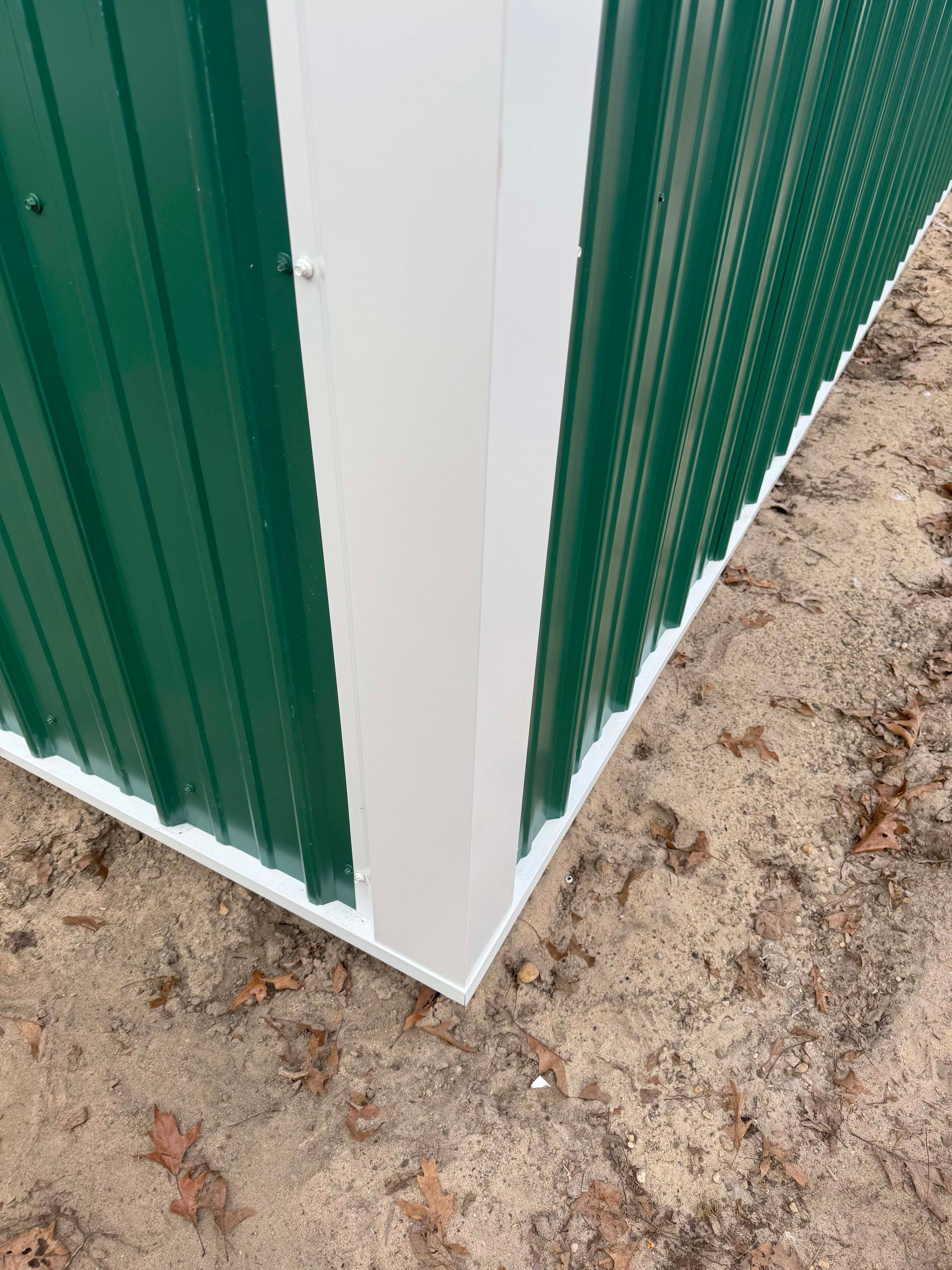 Green corrugated metal building corner with white trim, on dirt ground.
