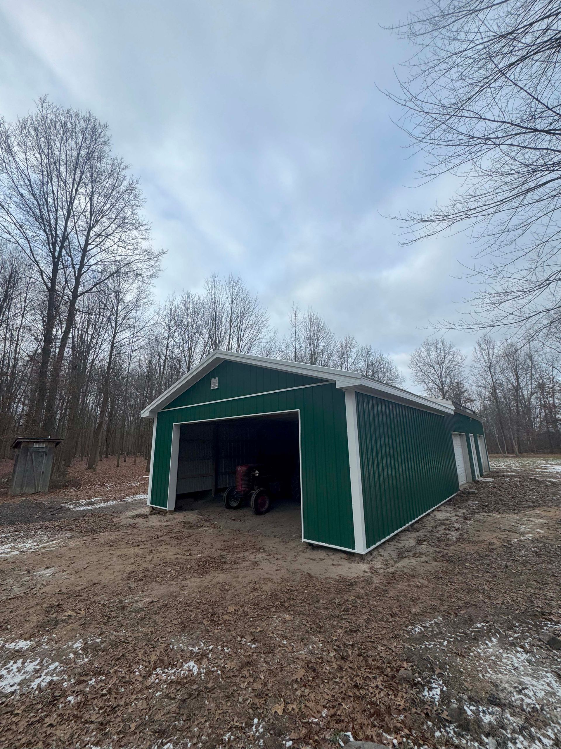 Green metal shed with open door, parked tractor inside; set in a wooded area under a cloudy sky.