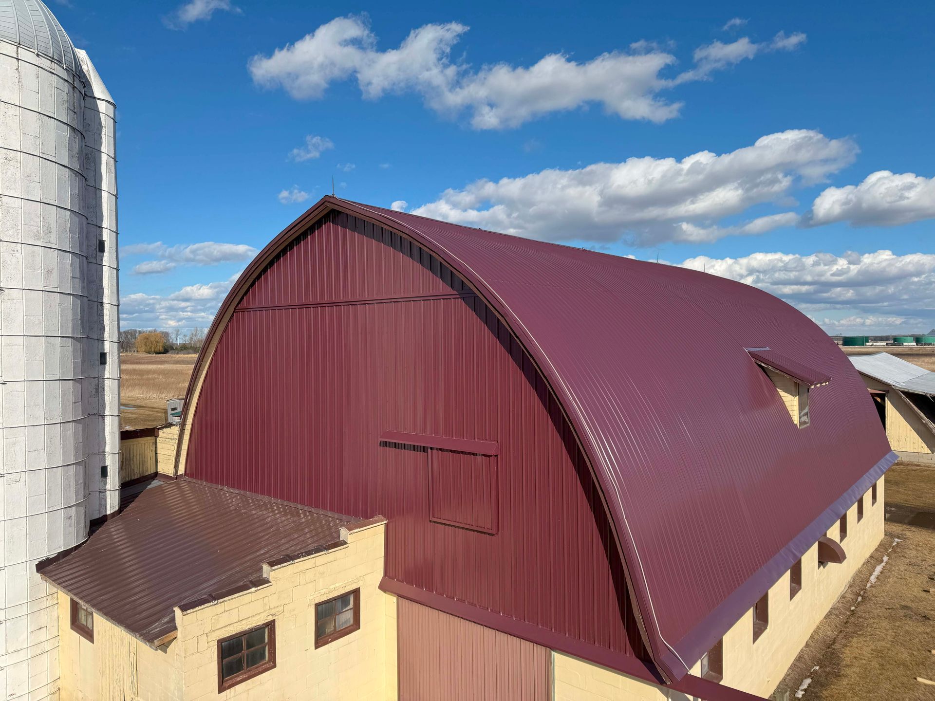 Barn with arched maroon roof and silo against a blue sky.