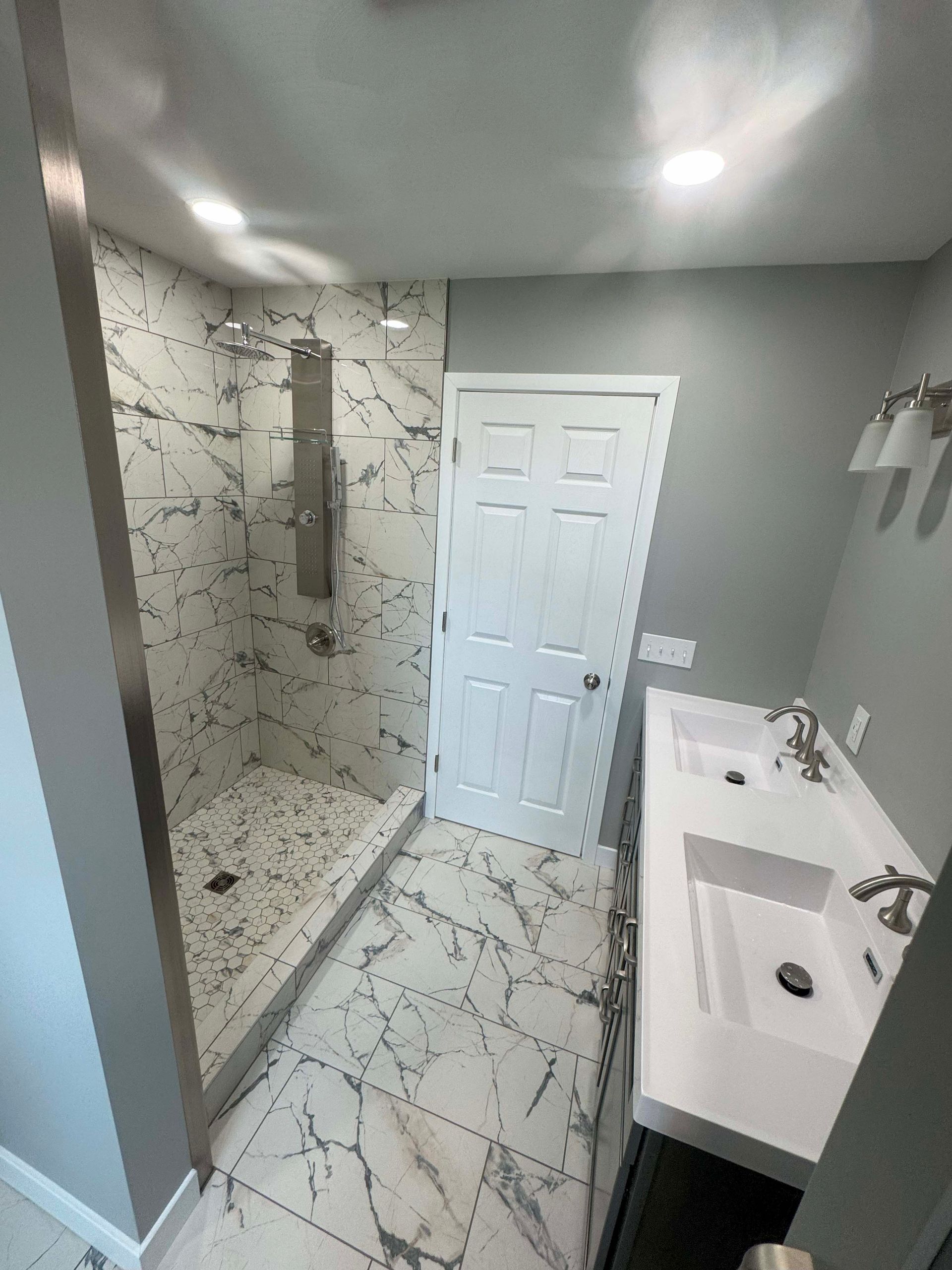 Modern bathroom with marble tile, glass shower, double sinks, and a white door.