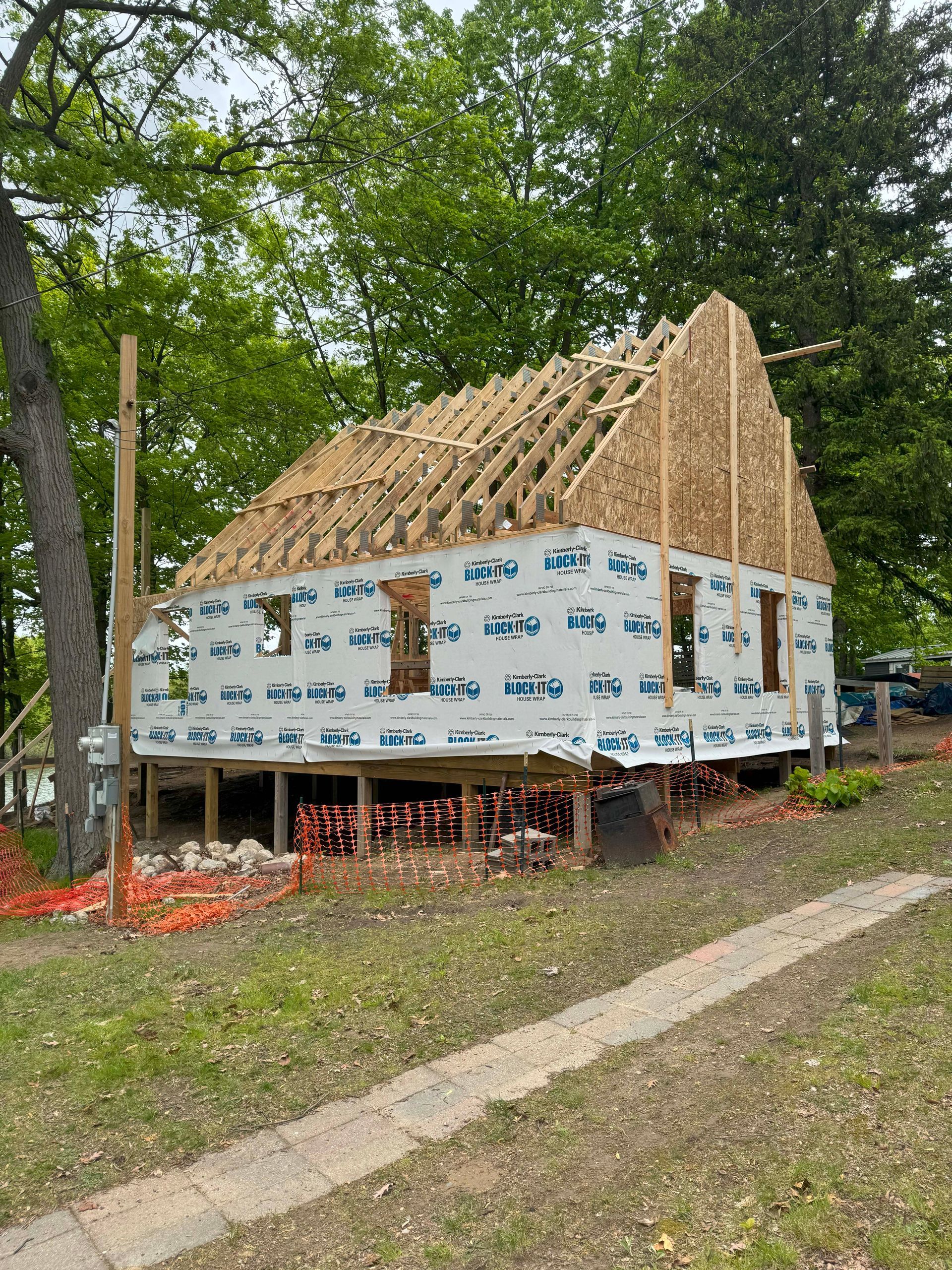 House under construction with exposed wooden frame and sheathing, blue wrap, in a wooded area.