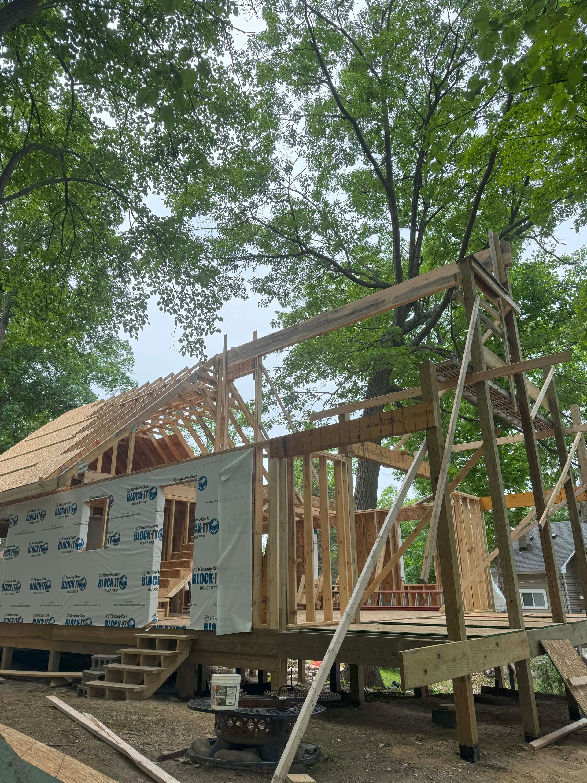 House under construction, wood framing visible. Deck with stairs, trees in background, cloudy sky.