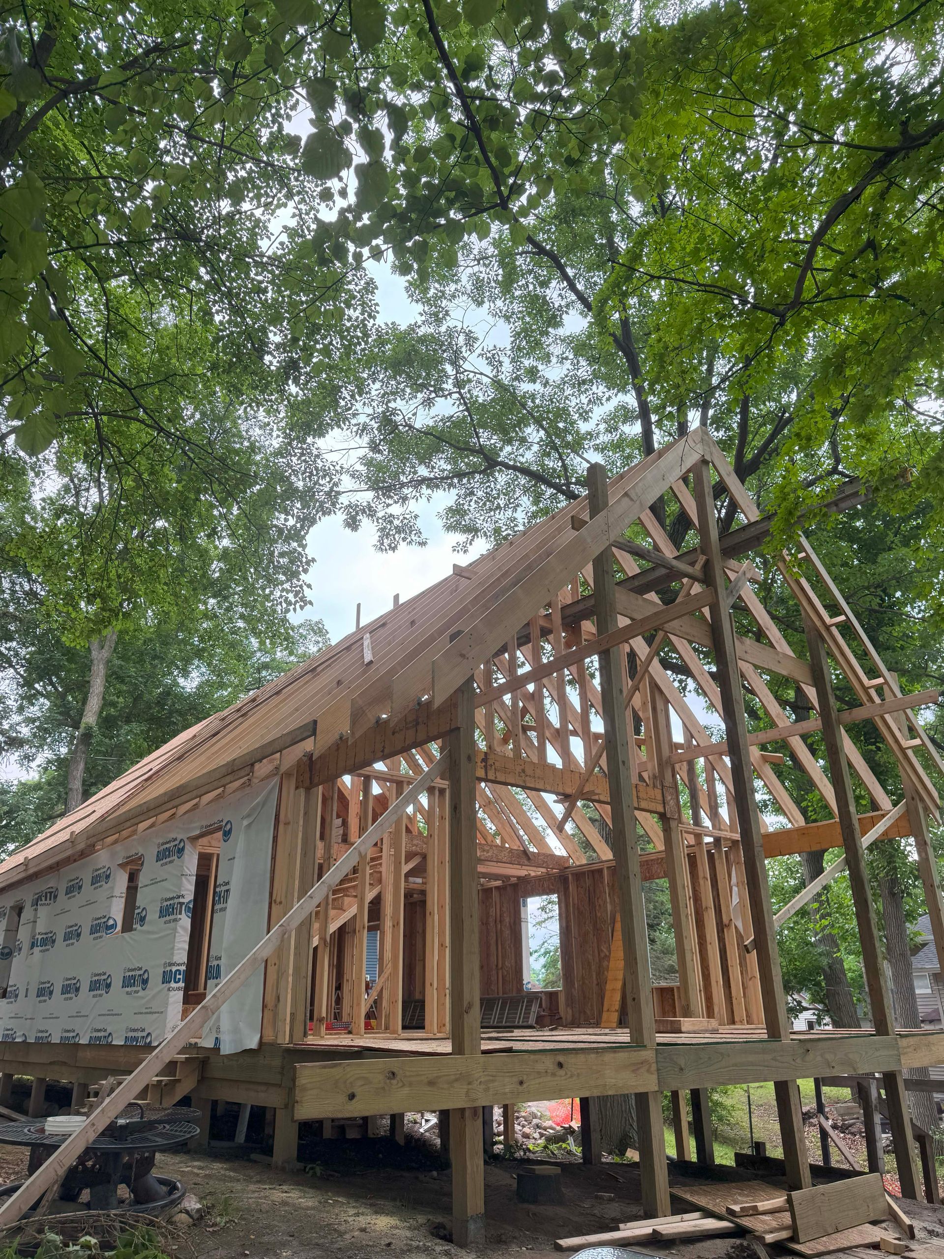 Wooden frame of a house under construction, surrounded by green trees and a blue sky.