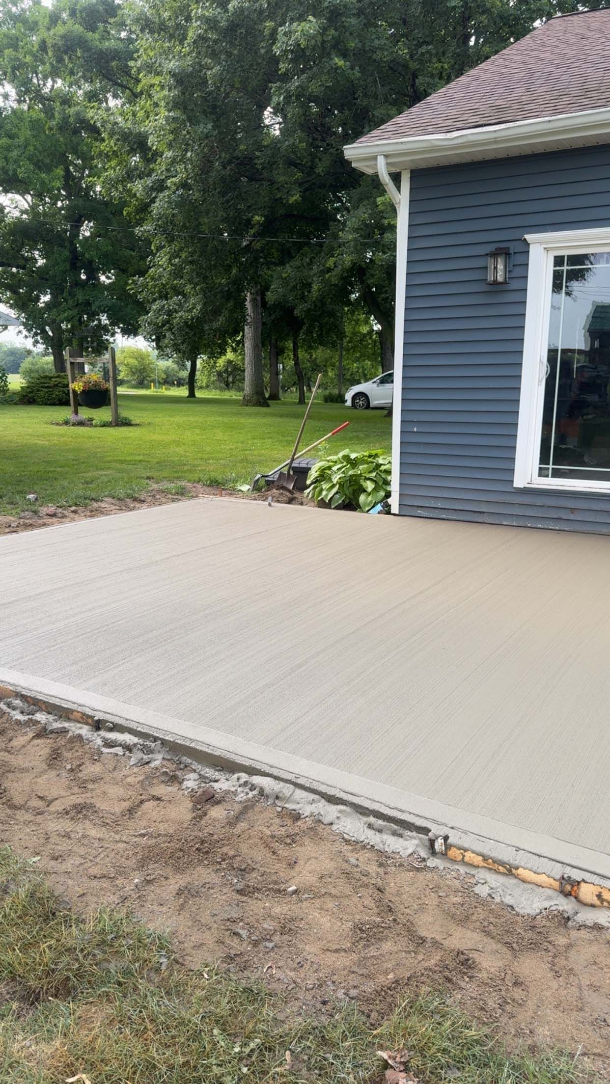 Newly poured concrete patio next to a blue-sided house, surrounded by grass and trees.