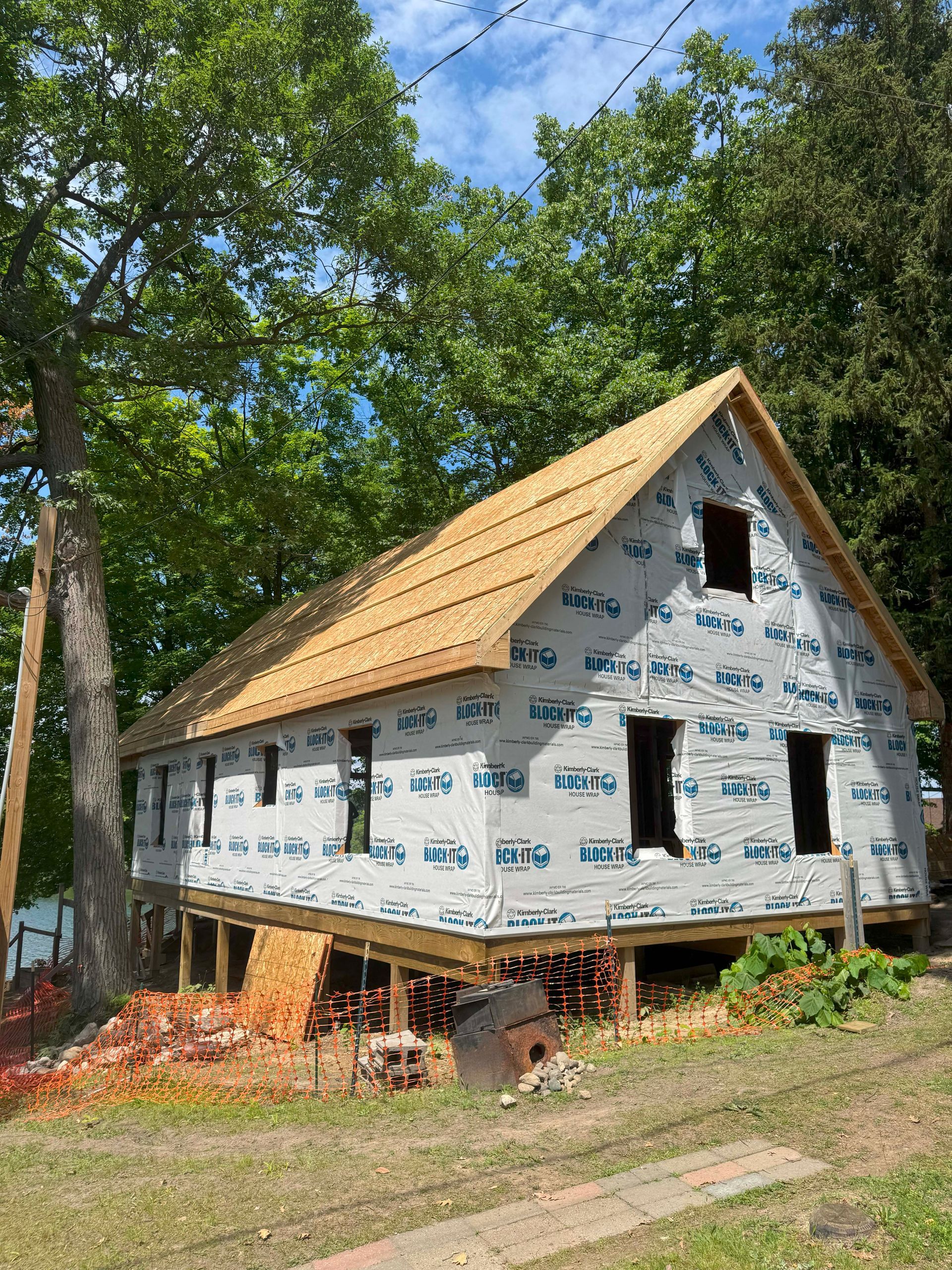Building under construction, wood frame with roof, windows, and protective wrap, near trees.