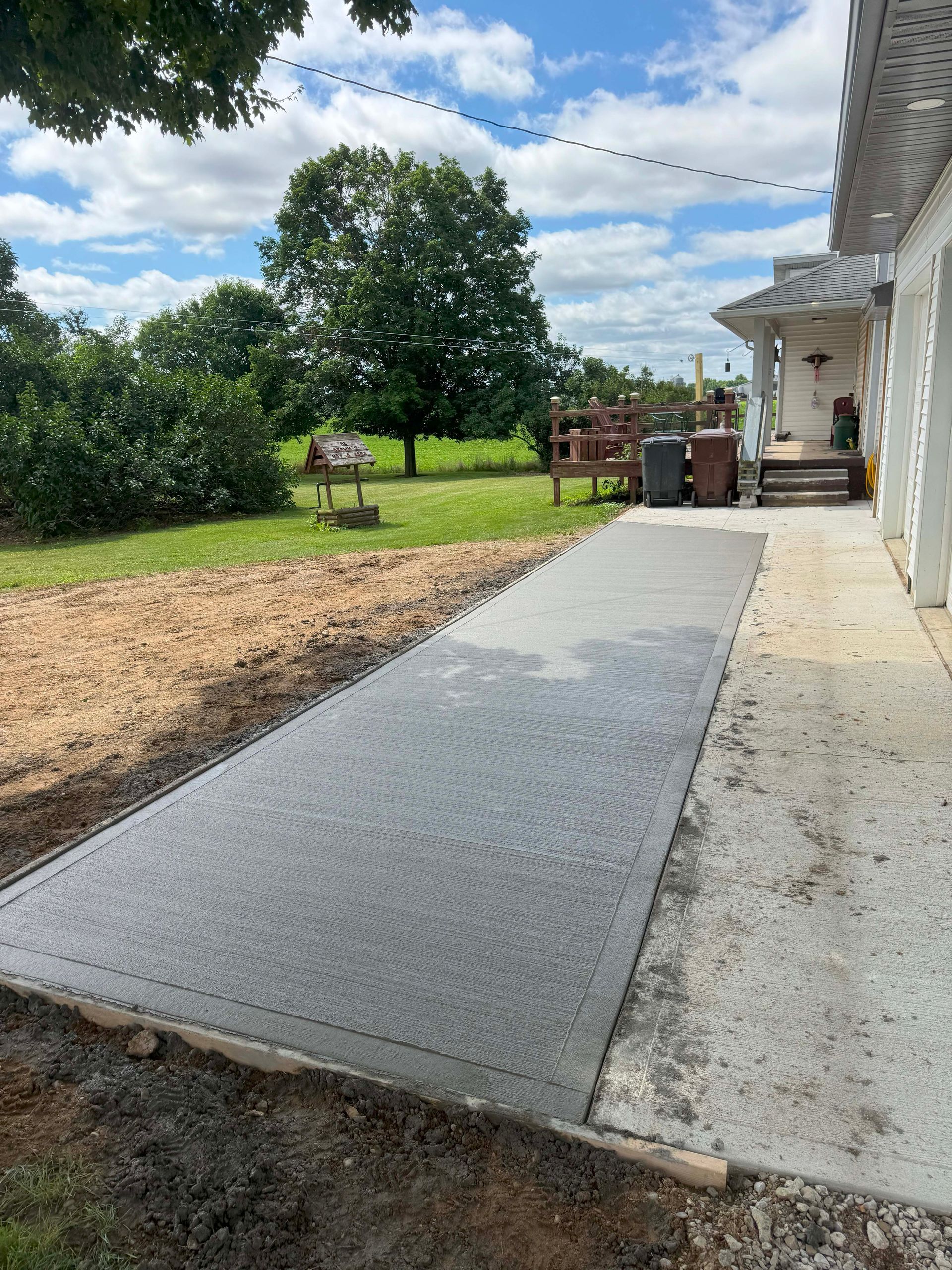 A concrete walkway next to a white house, leading to a patio, with grass and a tree in the background.