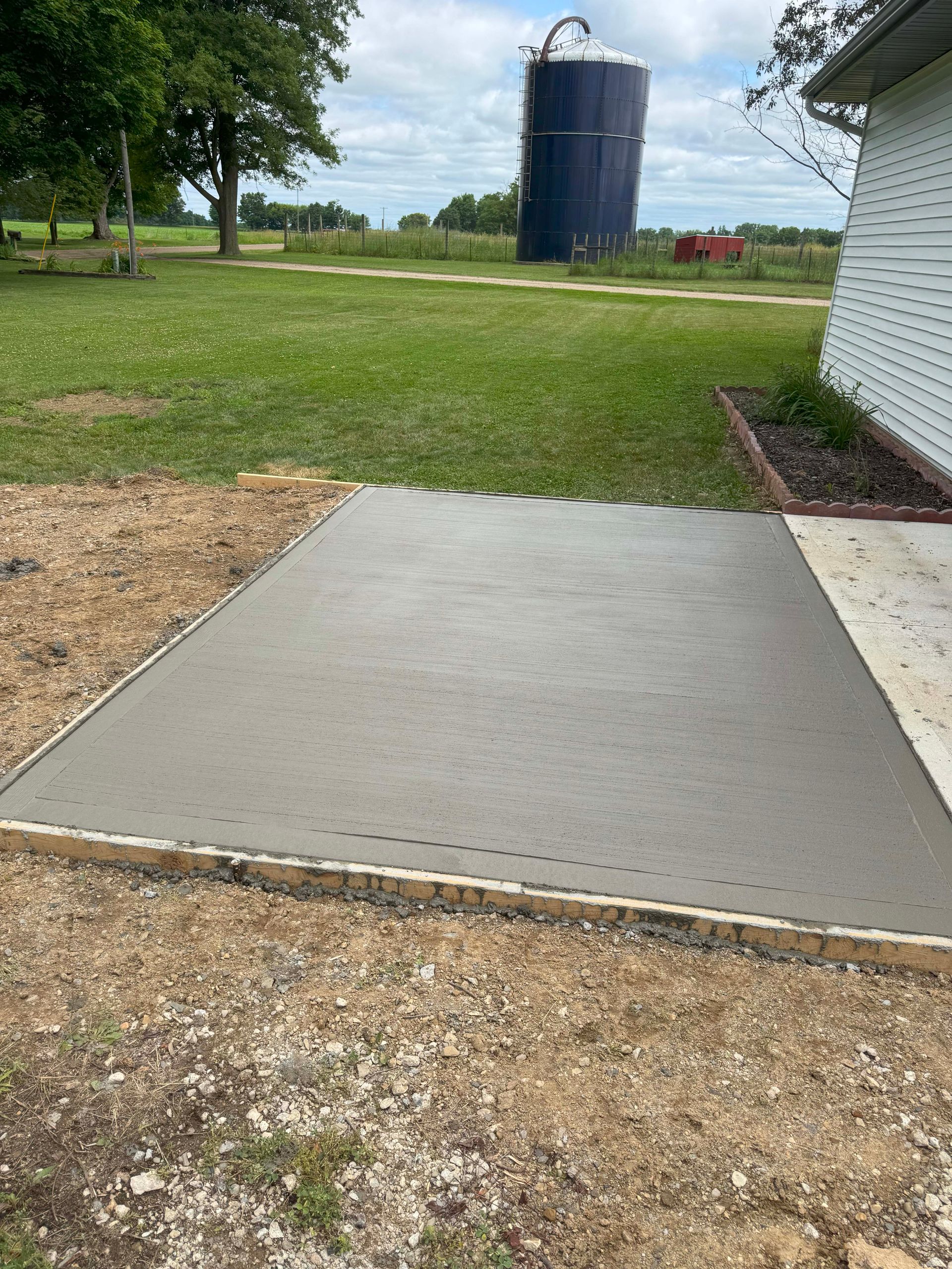 Newly poured concrete patio next to a house with a silo in the background.