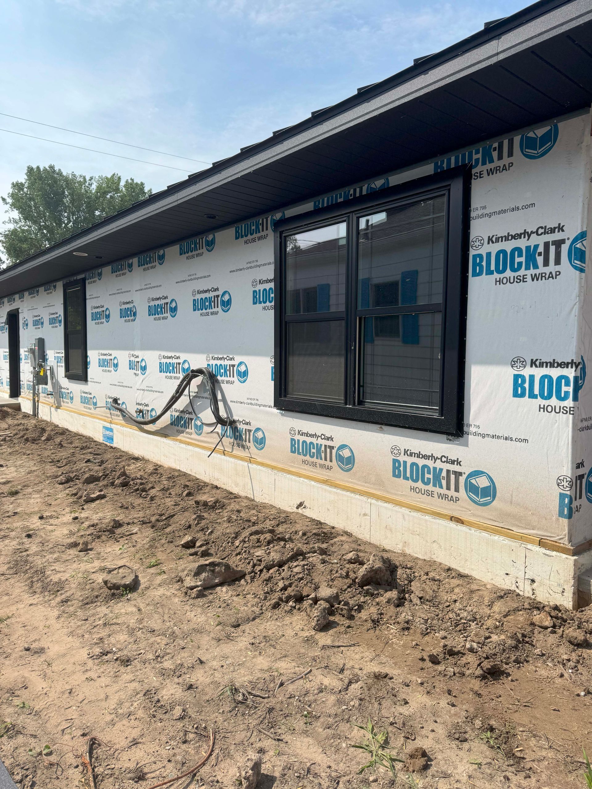 Exterior of a building under construction; black window frames, white protective wrap, brown soil in foreground.