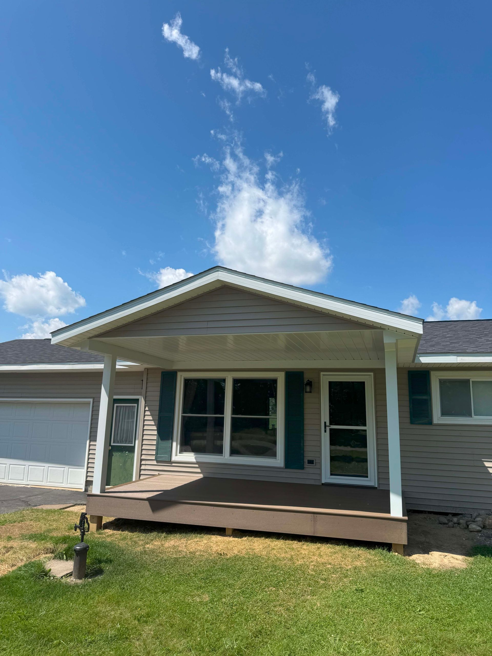 Ranch home with a porch, brown deck, and green shutters, under a partly cloudy blue sky.