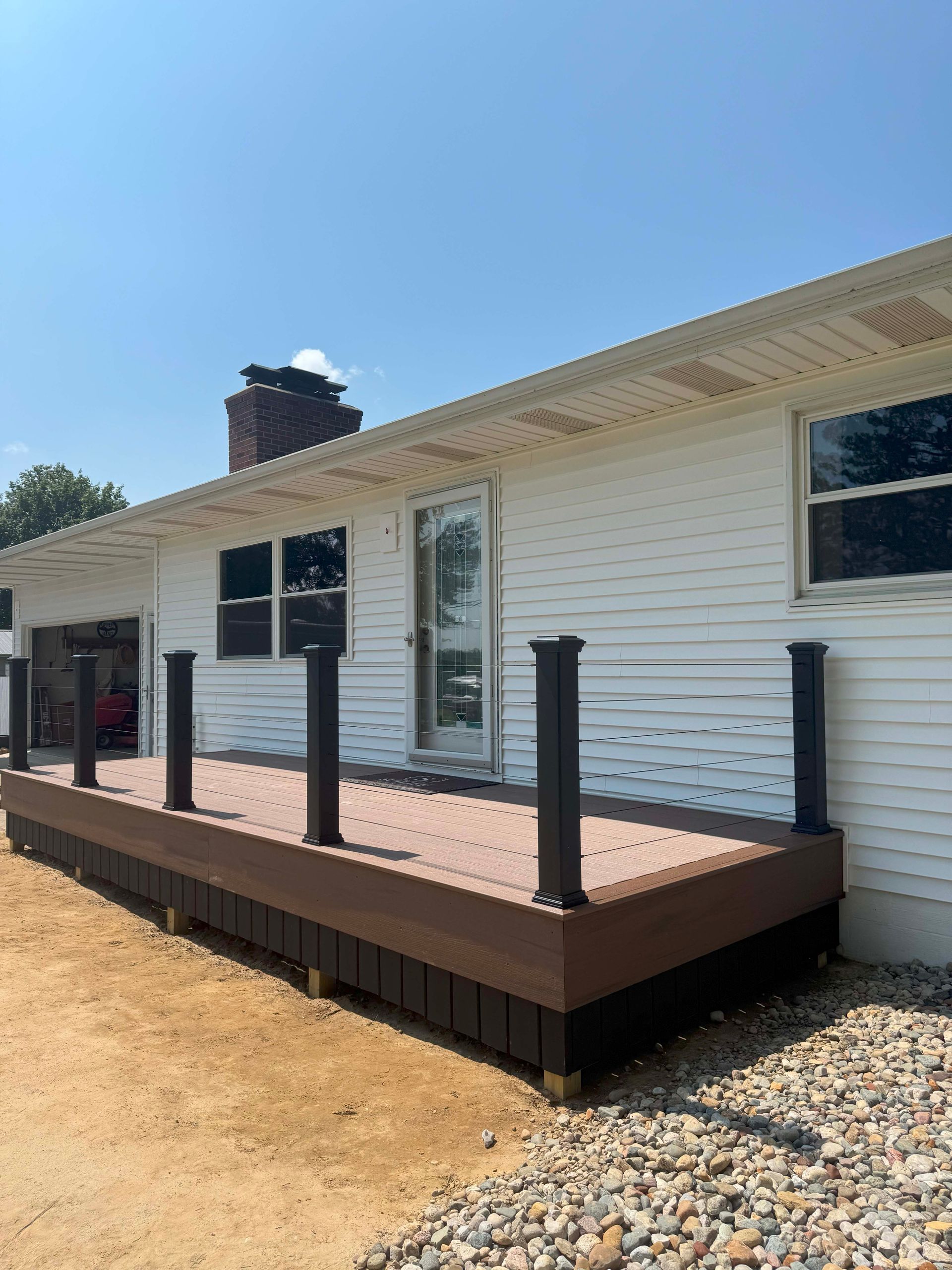Brown composite deck with black posts, attached to a white house with gravel ground.