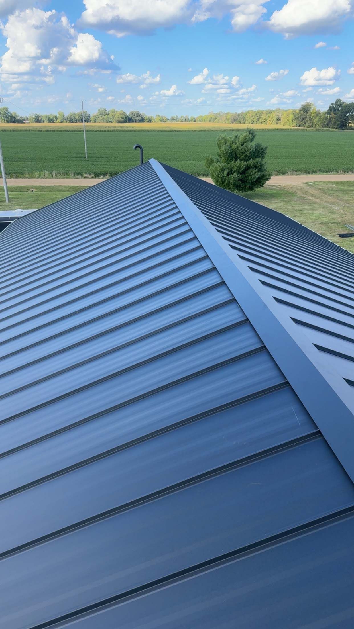 Metal roof with semicircular grooves, dark blue color. Visible in the background: fields and a blue sky with clouds.