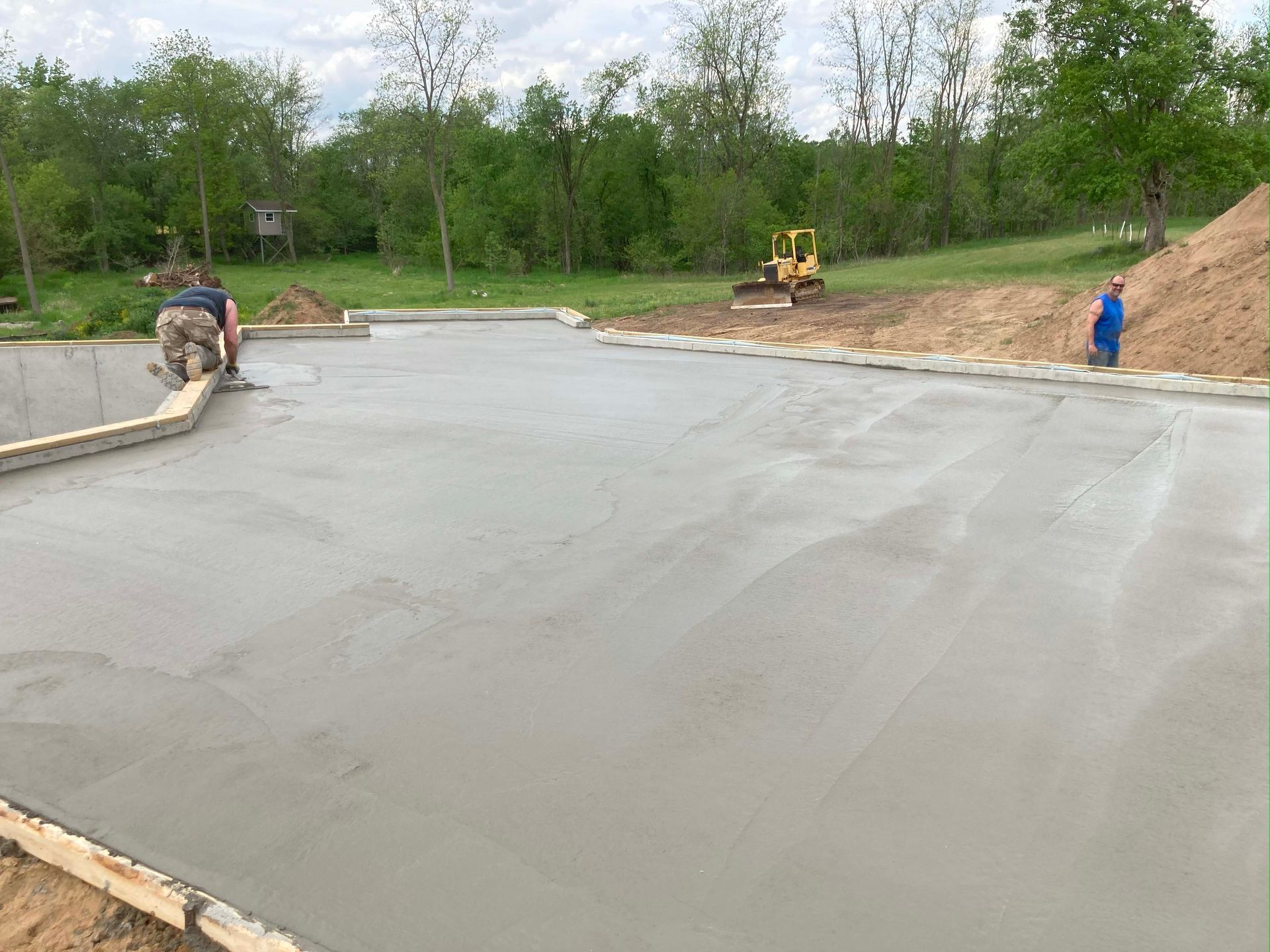 Workers smoothing wet concrete for a driveway in a rural setting; a small excavator is visible.
