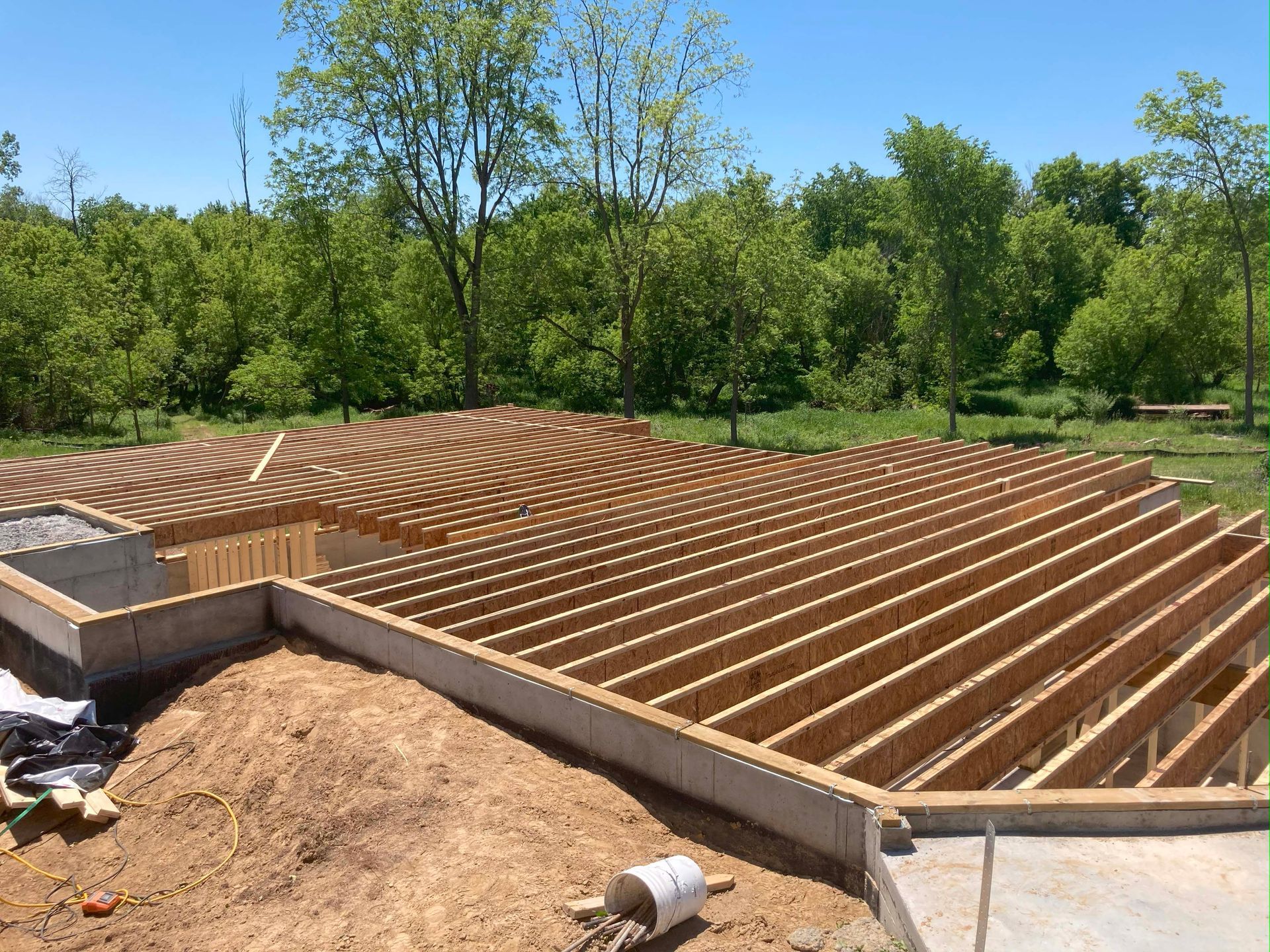 Wooden joists being laid over a foundation; construction site with a grassy, treed background.
