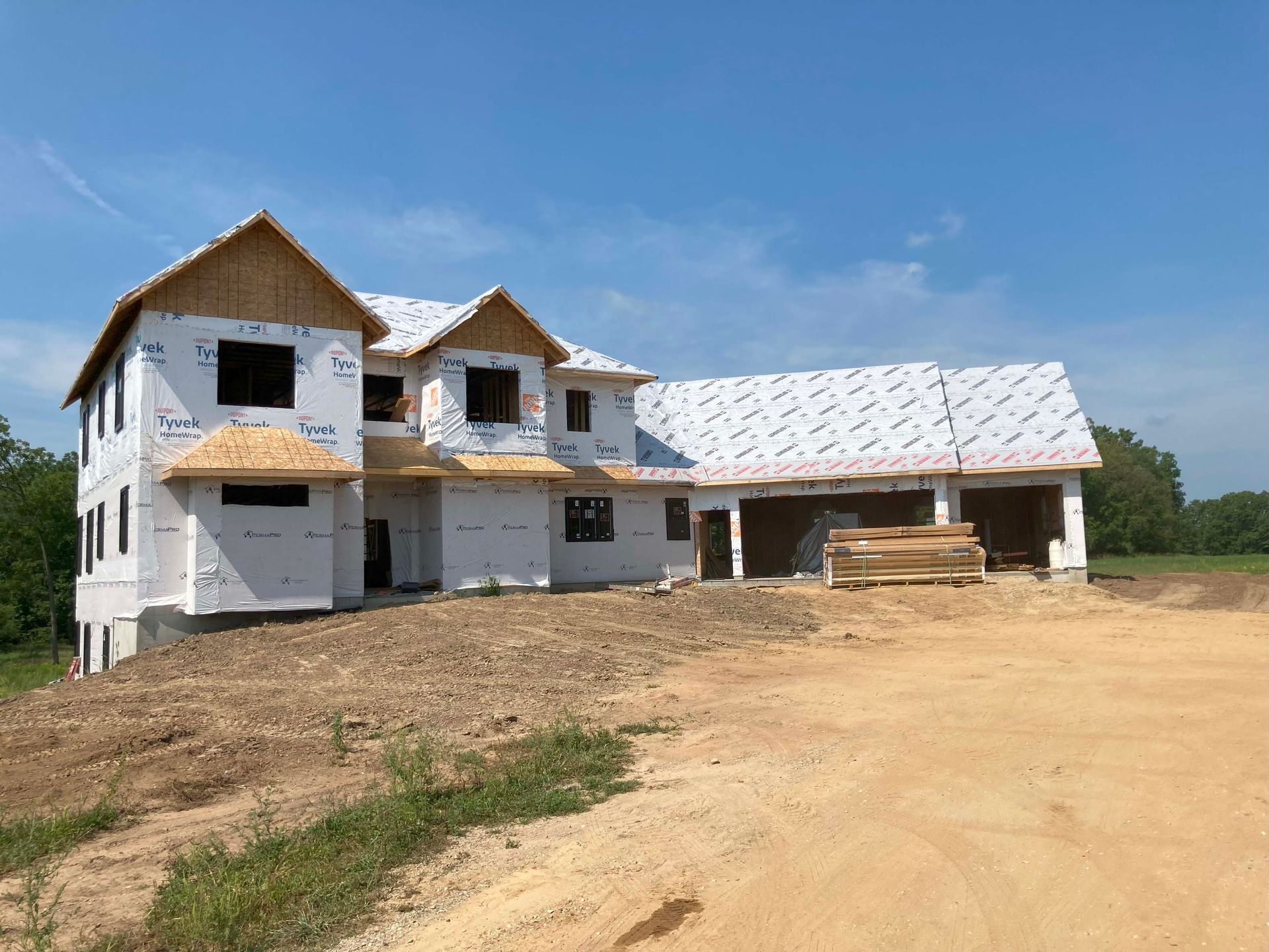 Two-story house under construction, blue sky, brown dirt, exposed wood roof supports and wrap.