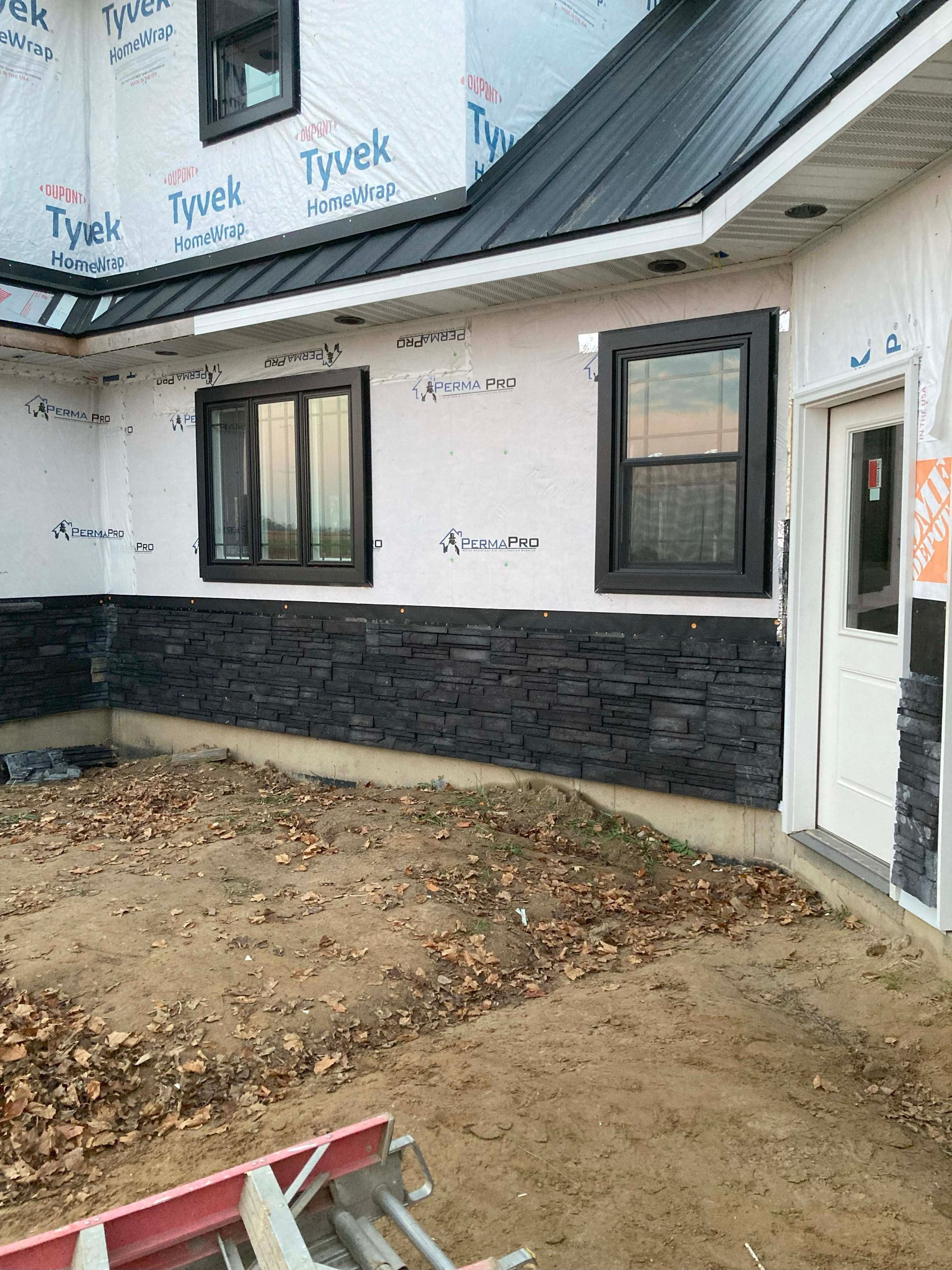 Exterior of a house under construction; black stone siding, black-framed windows, white door, Tyvek wrap, and sandy ground.