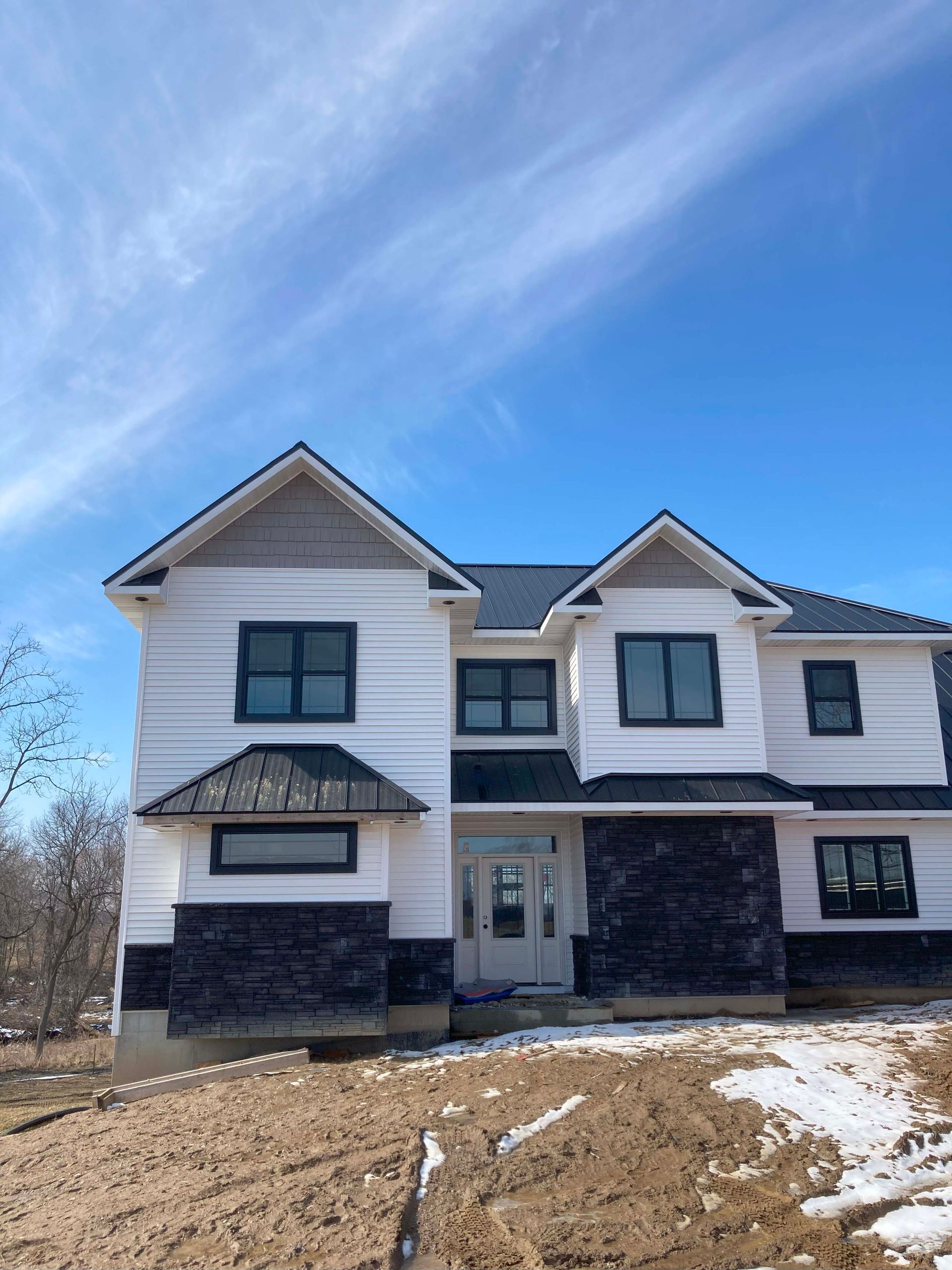 Two-story white house under construction with black trim and dark stone accent, blue sky.