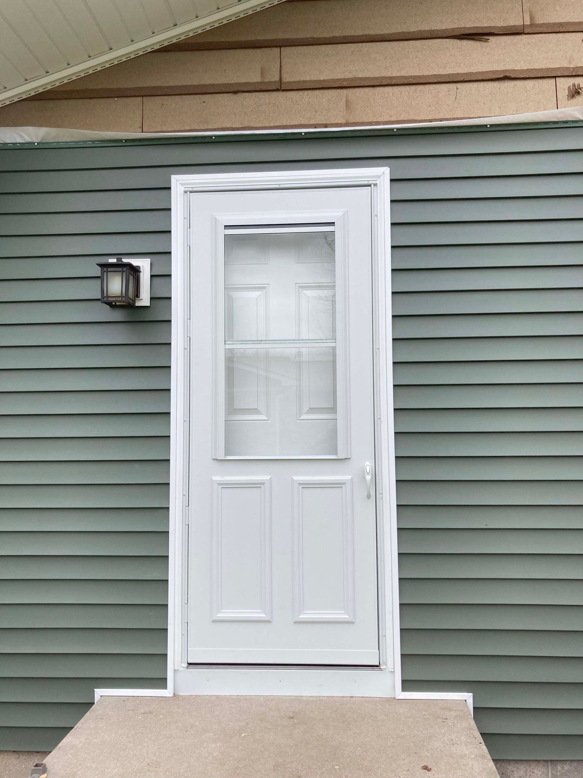 White door with glass panel, on green siding wall, with small porch and light fixture.