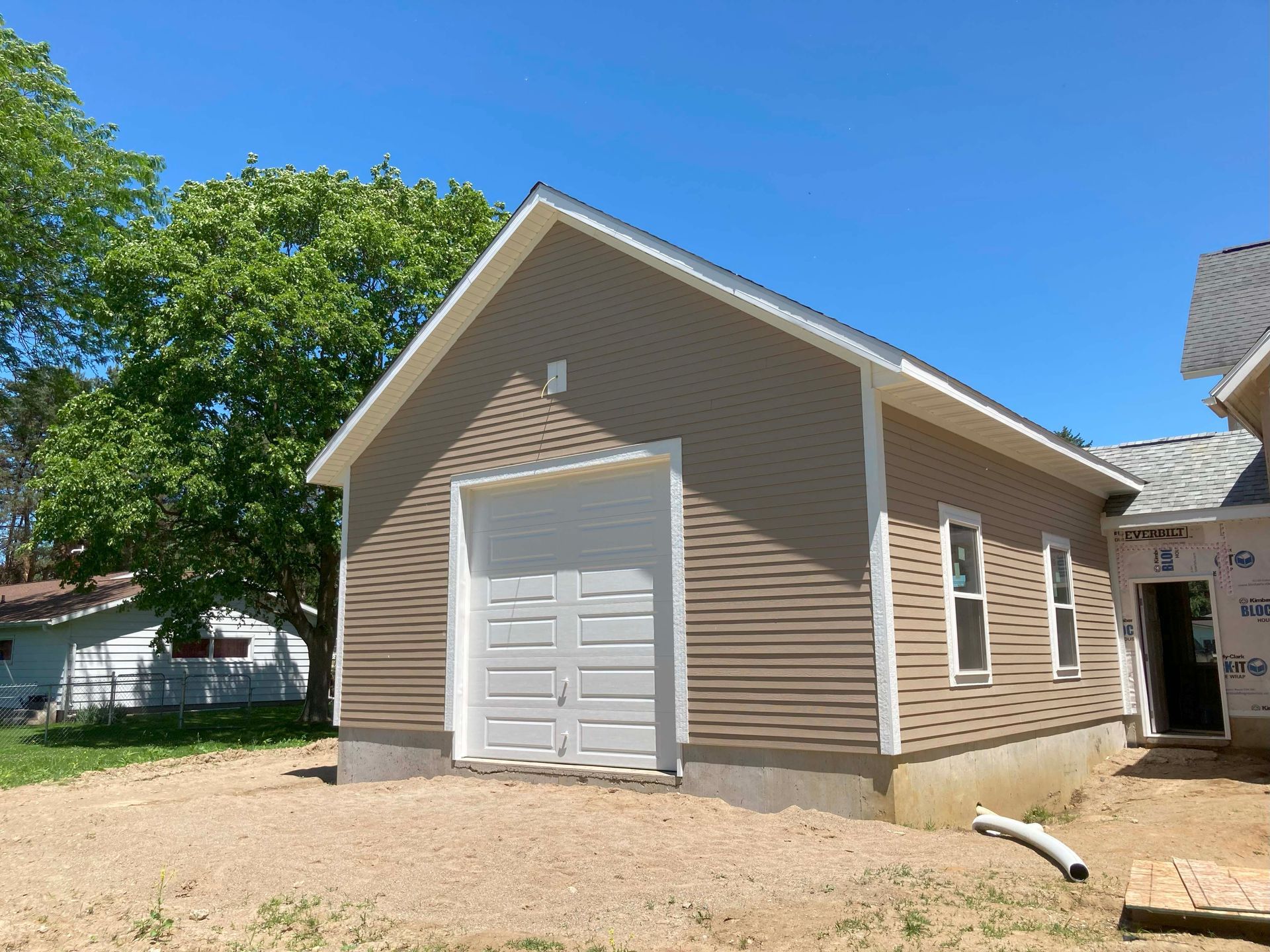 Tan-sided garage with white door and roof, under construction next to a house, on a sunny day.