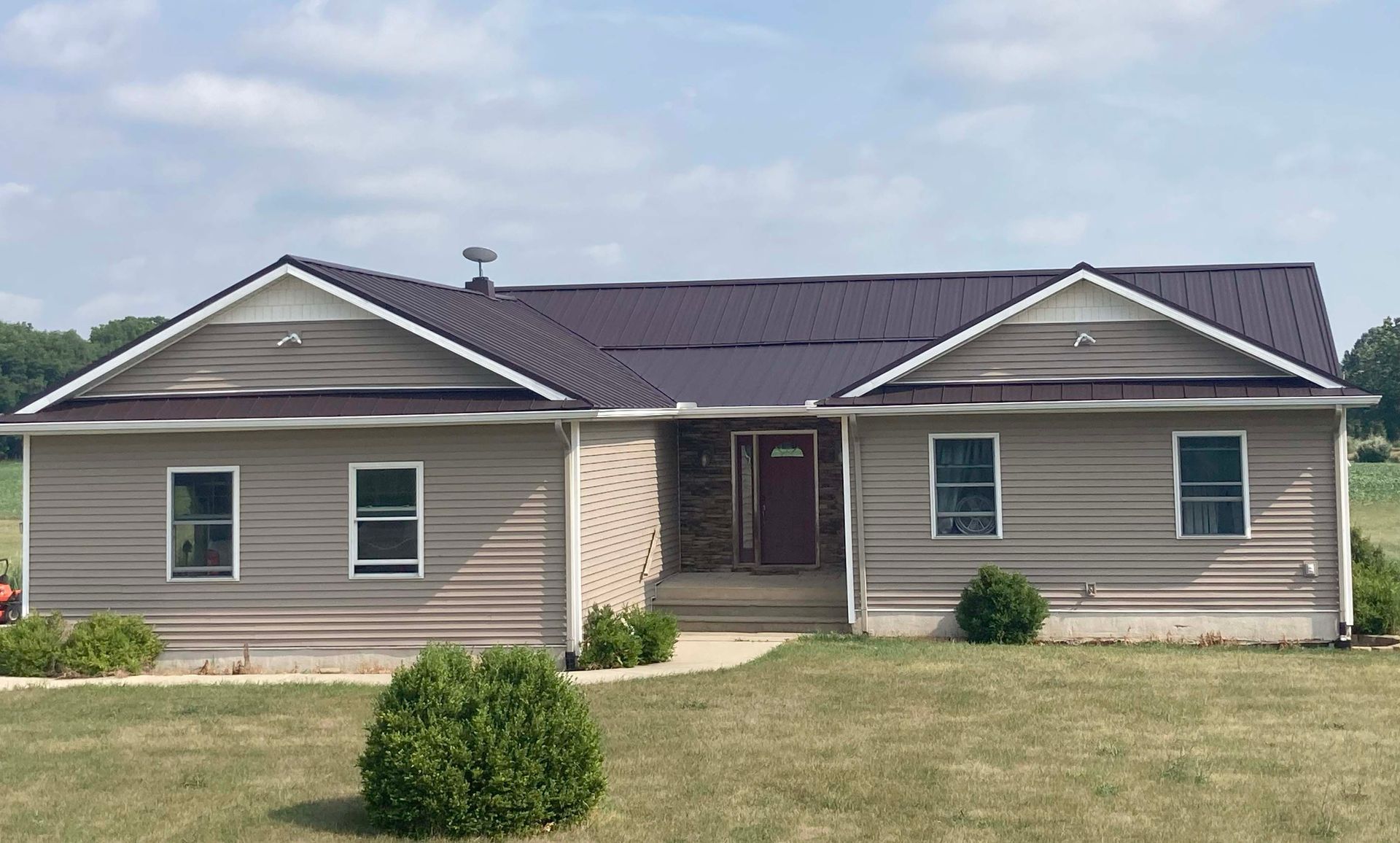 Tan house with a dark metal roof, three windows, and a stone entrance, set on a grassy lawn.