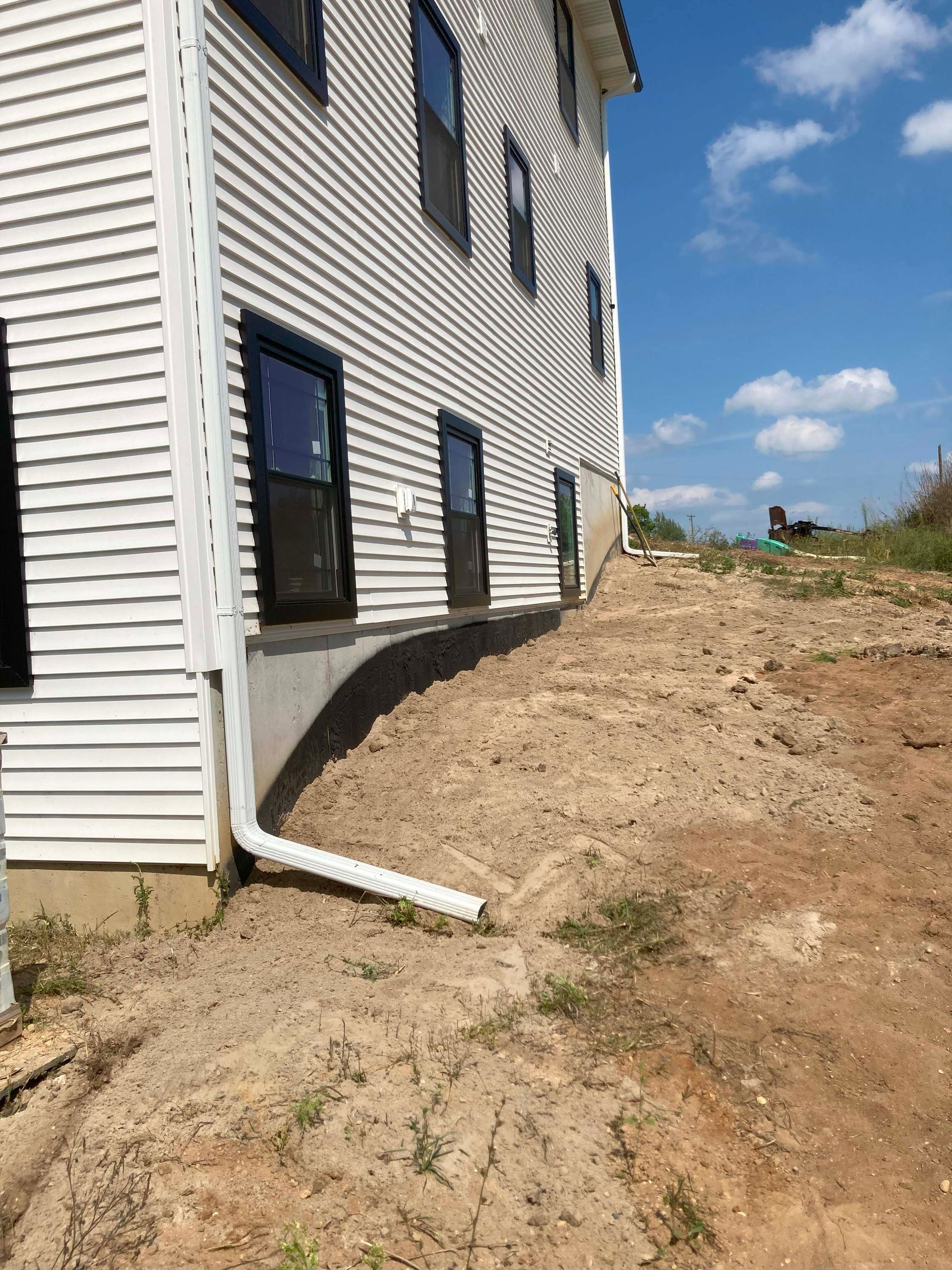 Side of a two-story building with white siding, black windows, and a dirt embankment.