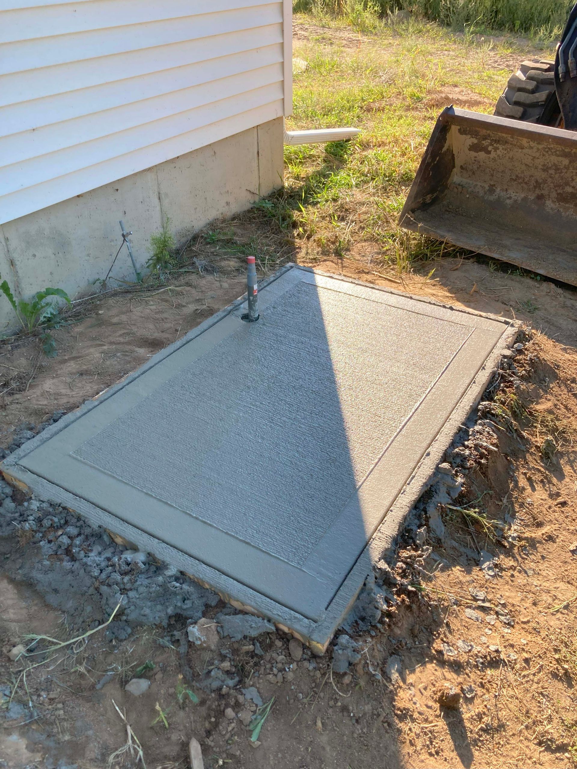 Concrete access cover in dirt near a white building, with a backhoe in the background.