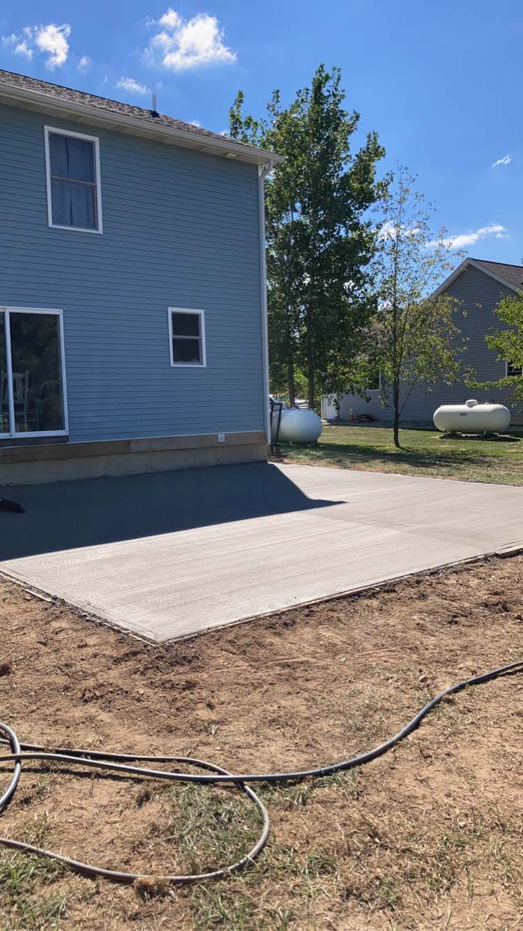 A new concrete patio next to a blue house, with a propane tank in the distance, on a sunny day.