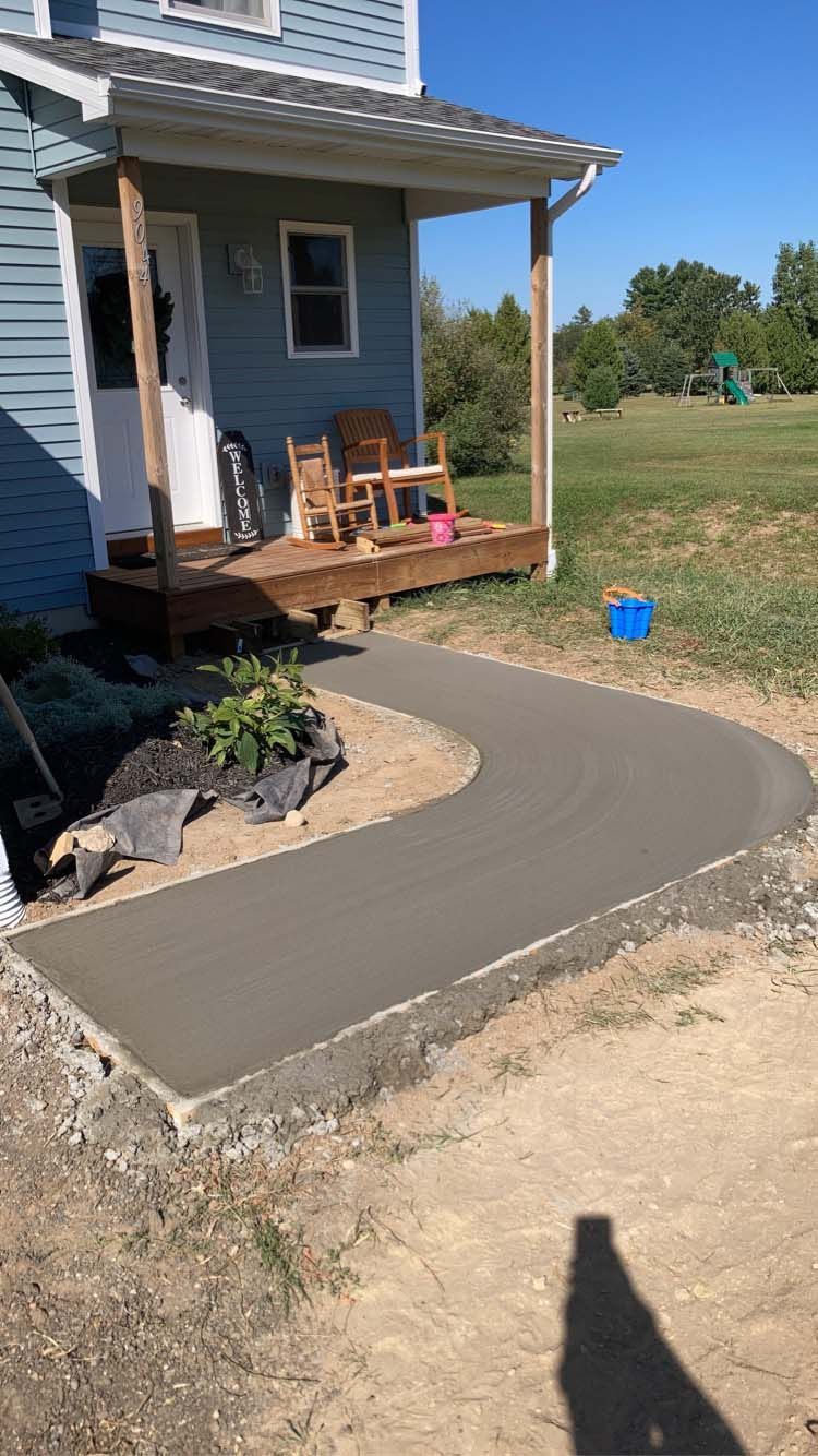 Newly poured concrete walkway curves toward a blue house with a porch; a grassy yard surrounds.