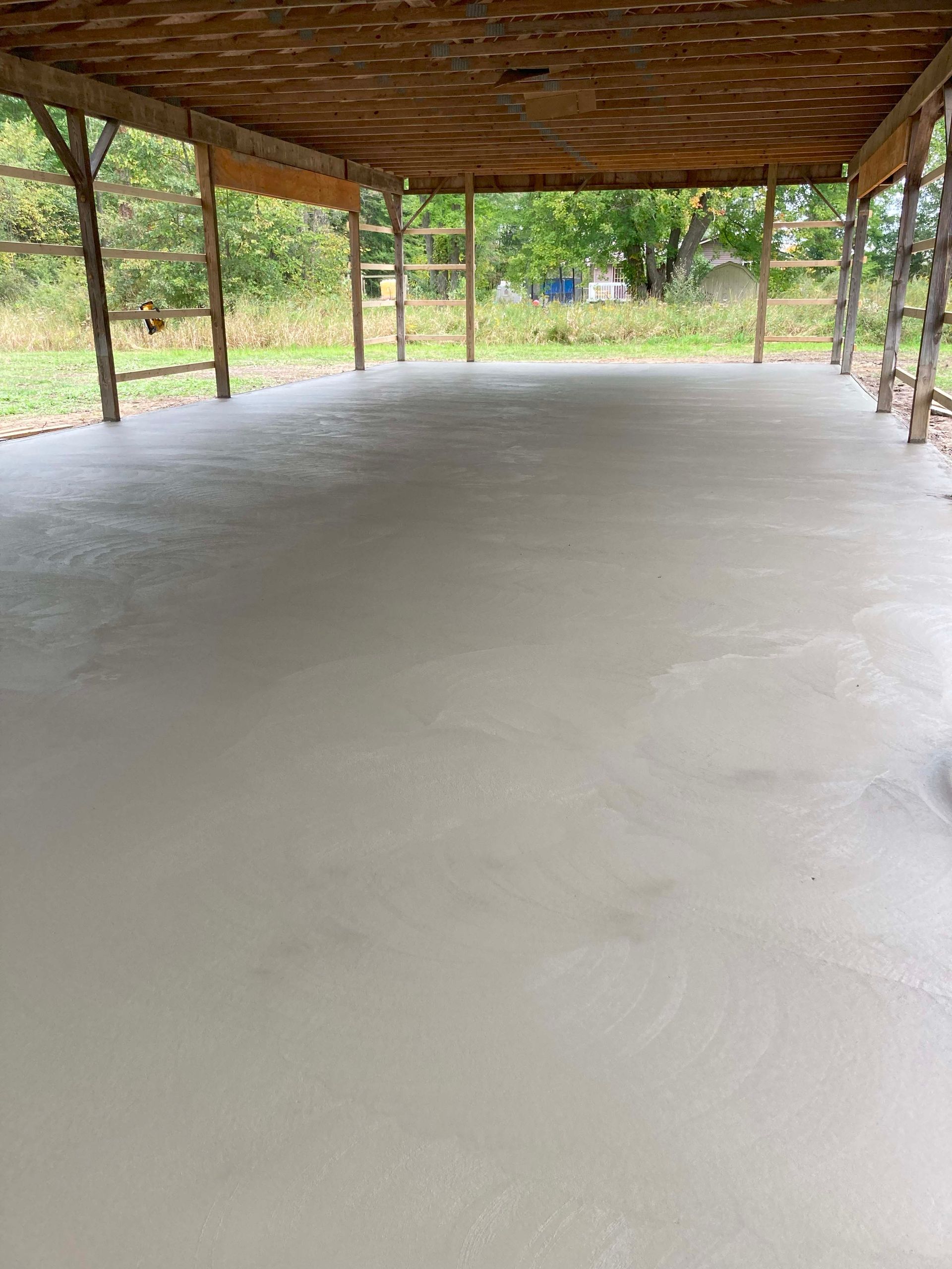 Newly poured concrete floor under a wooden roof structure, in a grassy outdoor area.