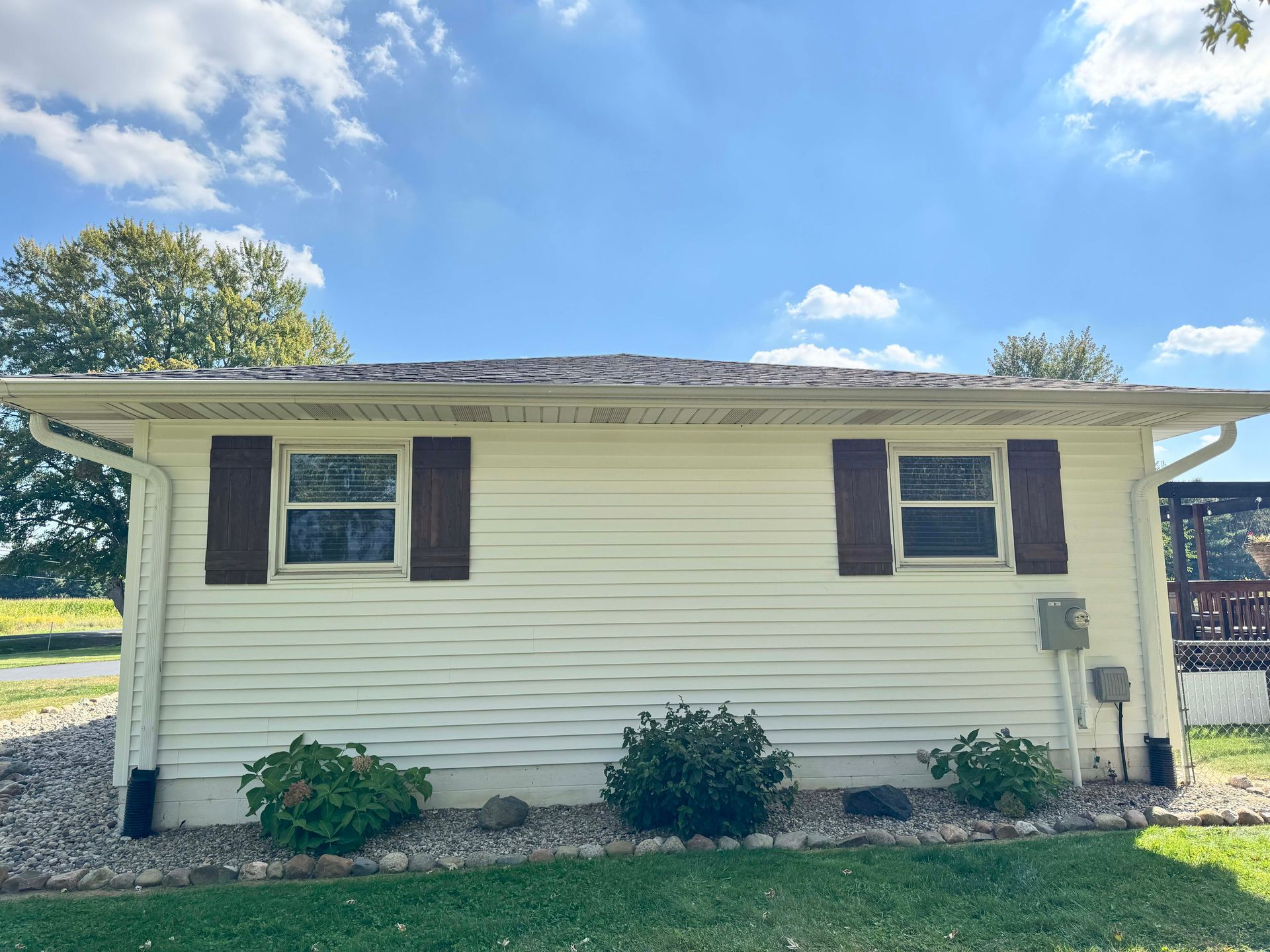 White-sided house with brown shutters, gutters, and roof. Blue sky with clouds.