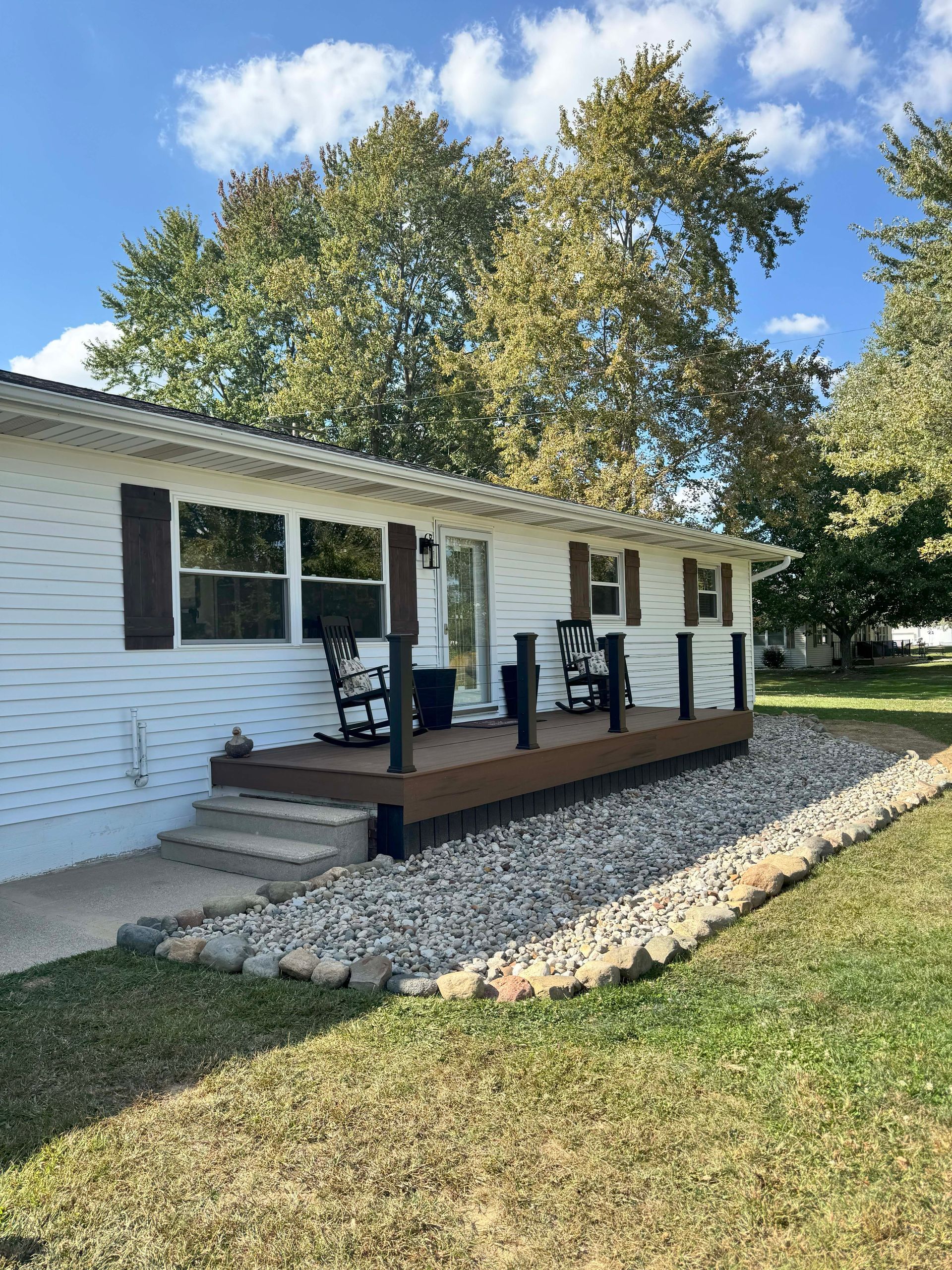 White house with brown shutters and deck, black railings, rock landscaping, and two rocking chairs.