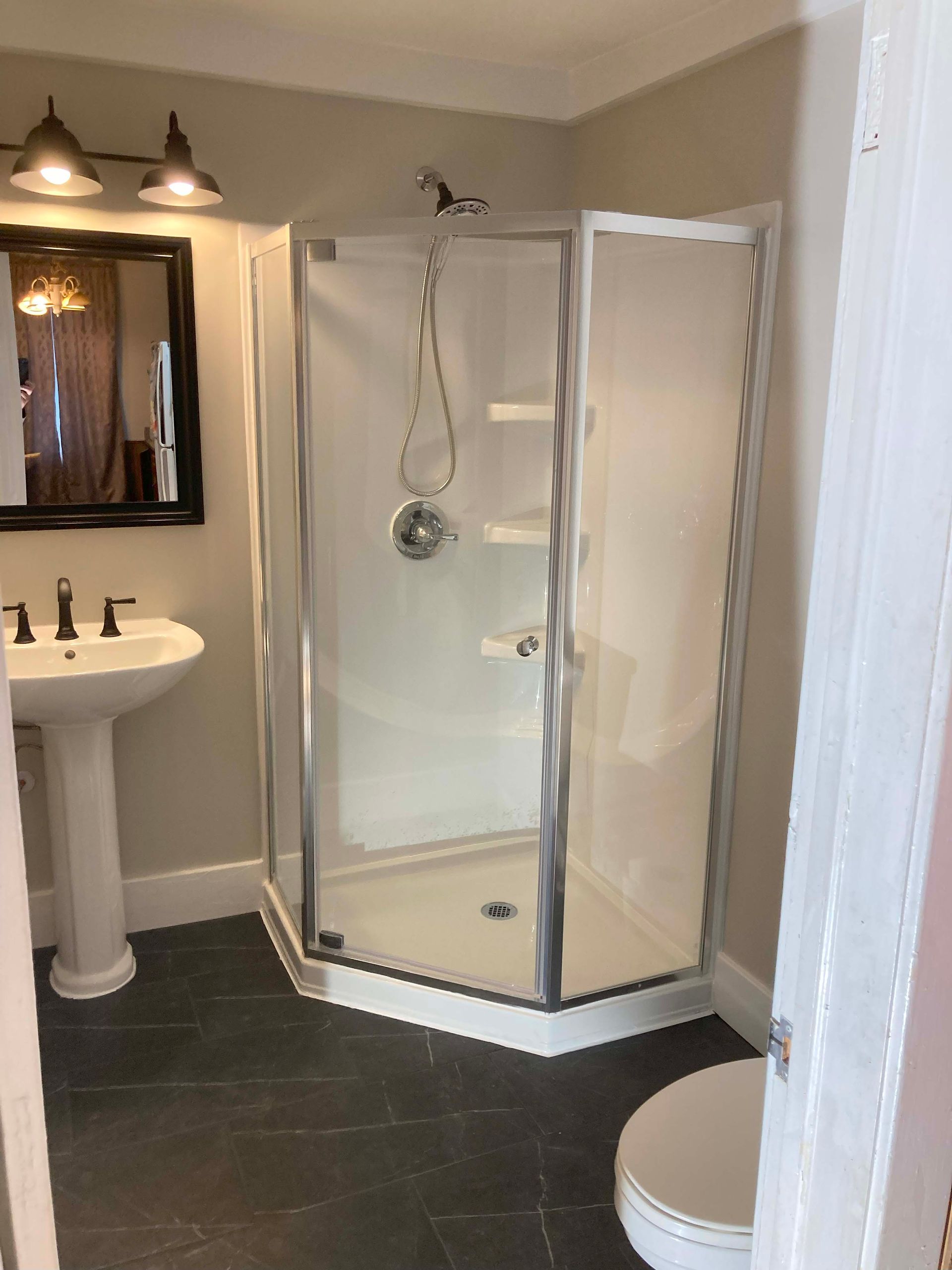 Bathroom with a corner shower, sink, mirror, and toilet; gray walls and dark tiled floor.
