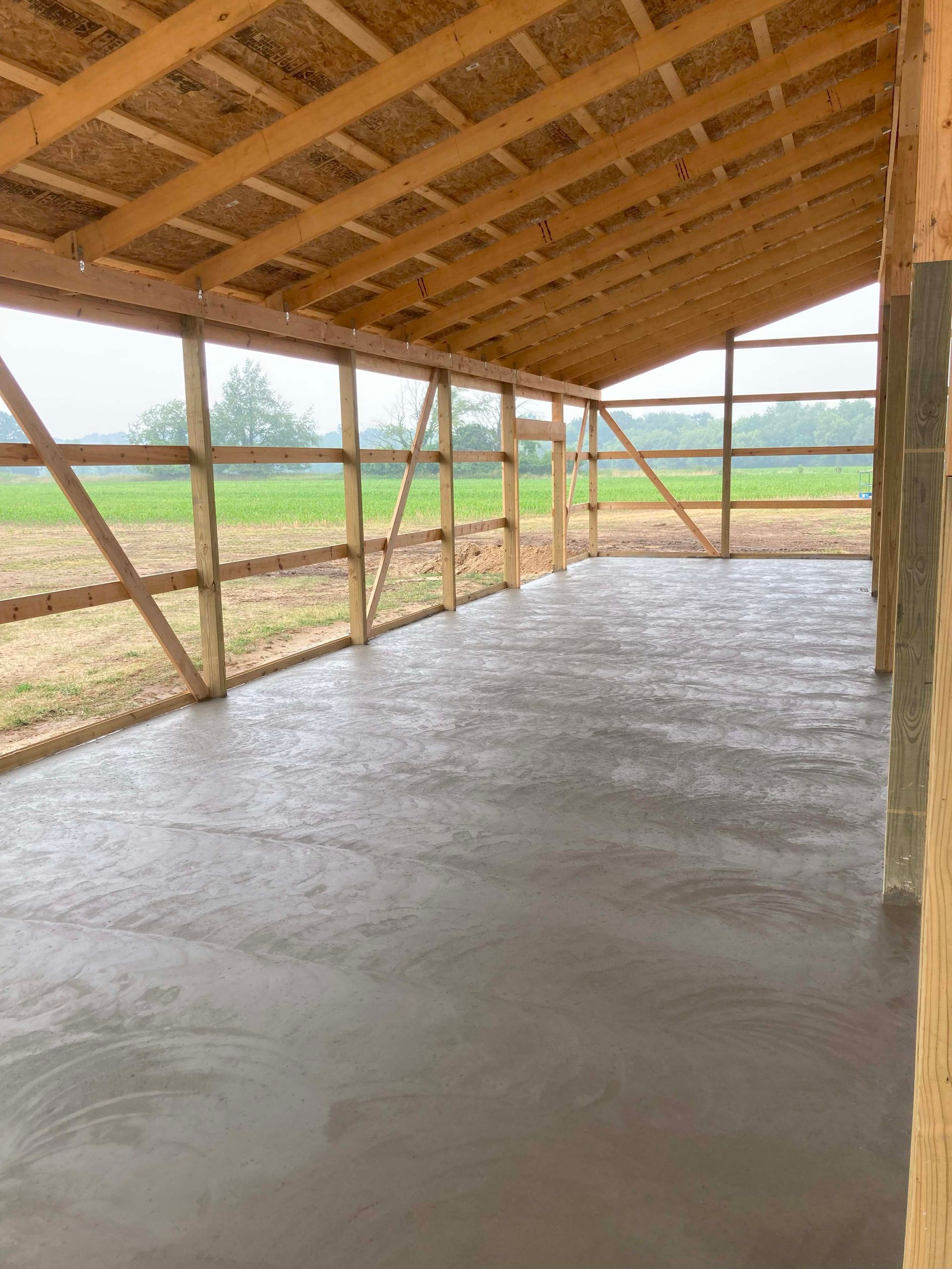 Newly poured concrete floor inside a wooden structure with an open side, overlooking a field.