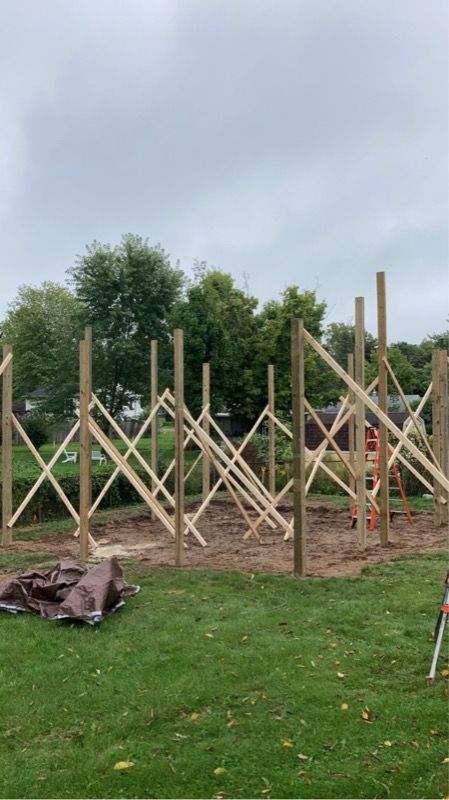 Construction of a wooden frame structure in a yard. Posts and cross bracing visible under a cloudy sky.